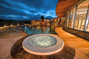 A hot tub with a wooden deck, a pool, and a building with large windows and a stone facade. - Hotel Atlantis (Rozdrojovice)