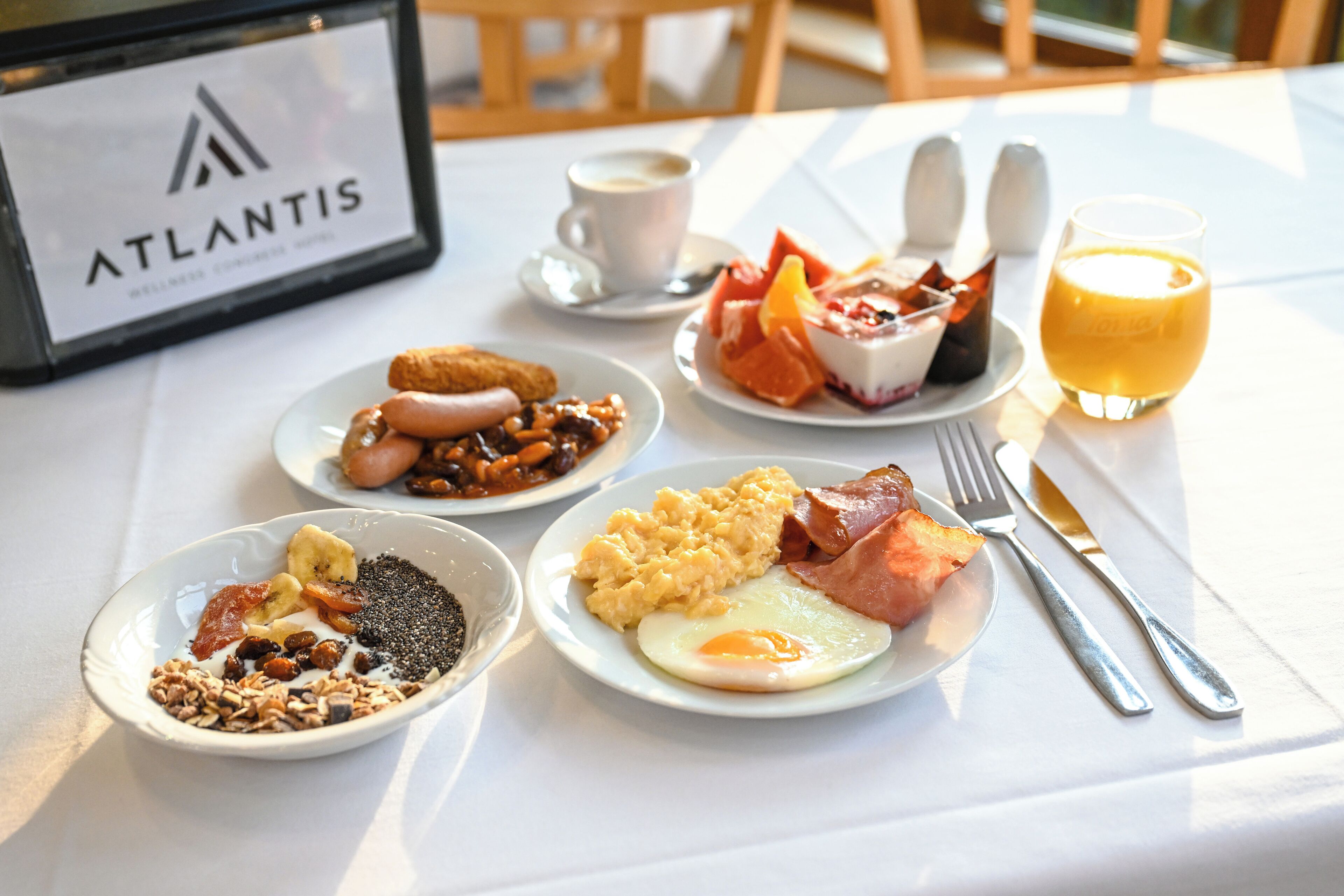 Desayuno con platos de huevos, tocino y fruta, una taza de café y un vaso de jugo sobre una mesa con un letrero que dice 'Atlantis'.