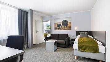 Une chambre d’hôtel moderne avec un canapé gris, un lit blanc, une petite table blanche et une grande fenêtre avec des rideaux.