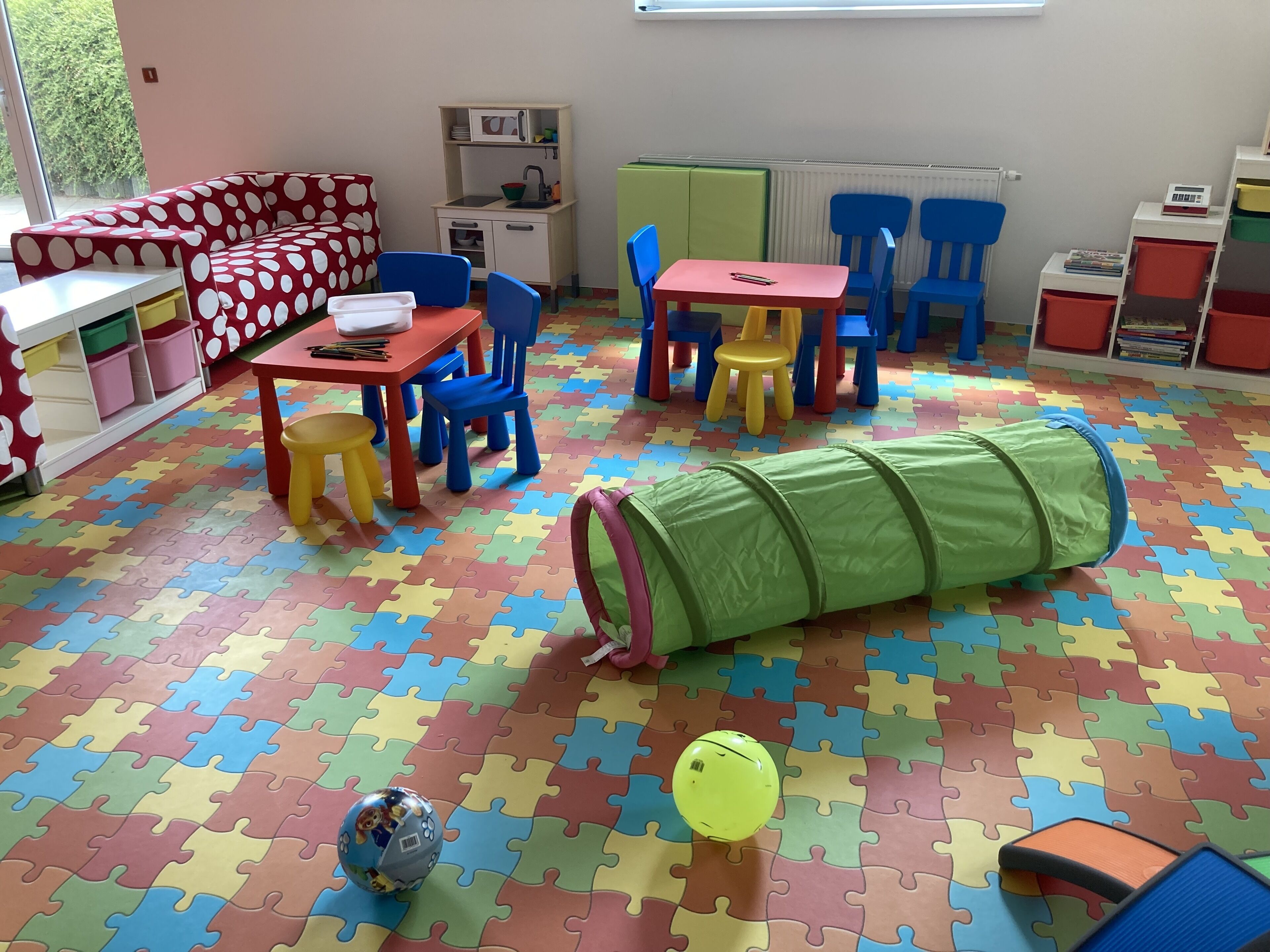 Una sala de juegos con piso de baldosas tipo rompecabezas, un sofá, una mesa, sillas, una pequeña zona de cocina y un túnel verde.