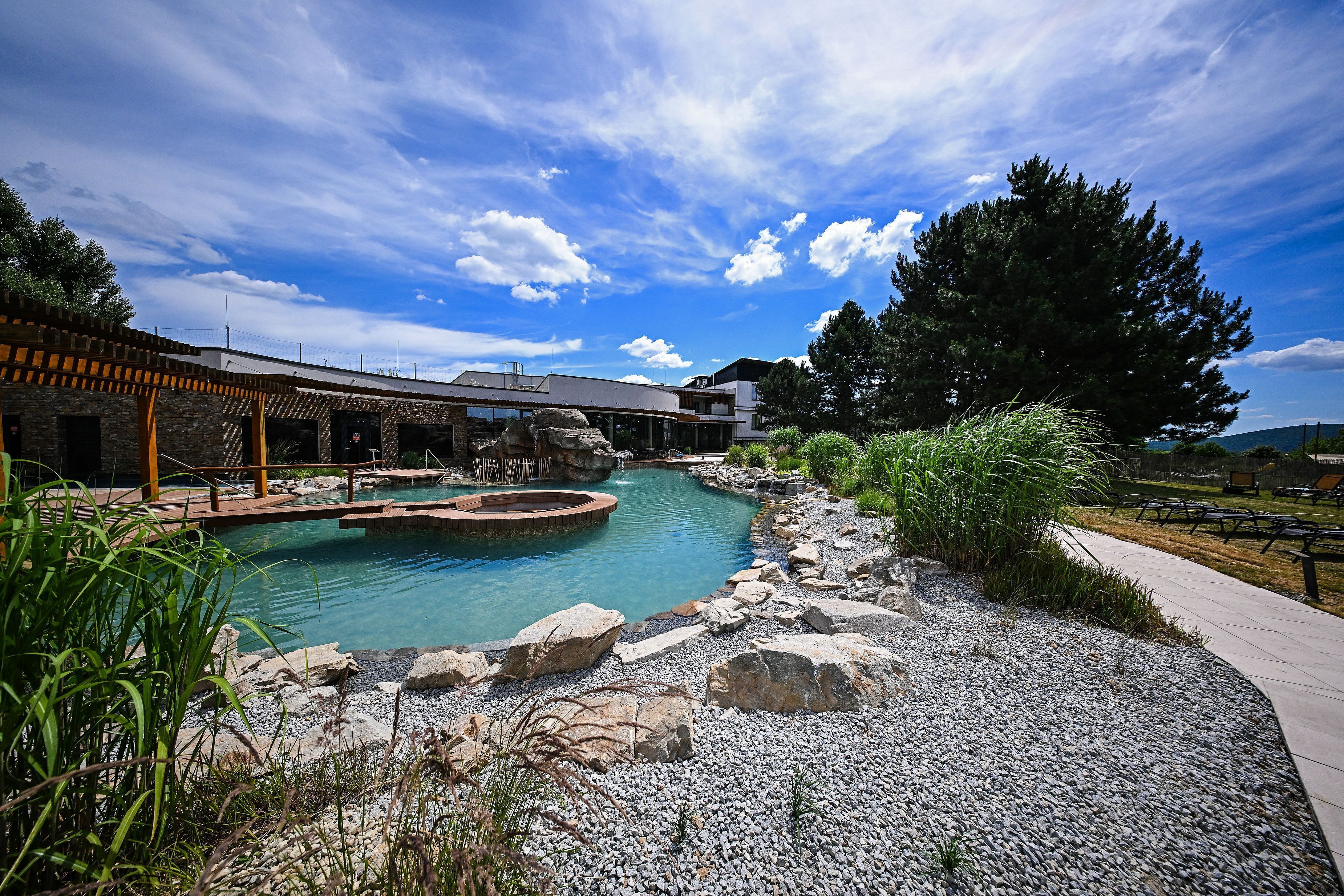 Zona de piscina con terraza de piedra, un pérgola de madera y vistas a un edificio de diseño moderno.