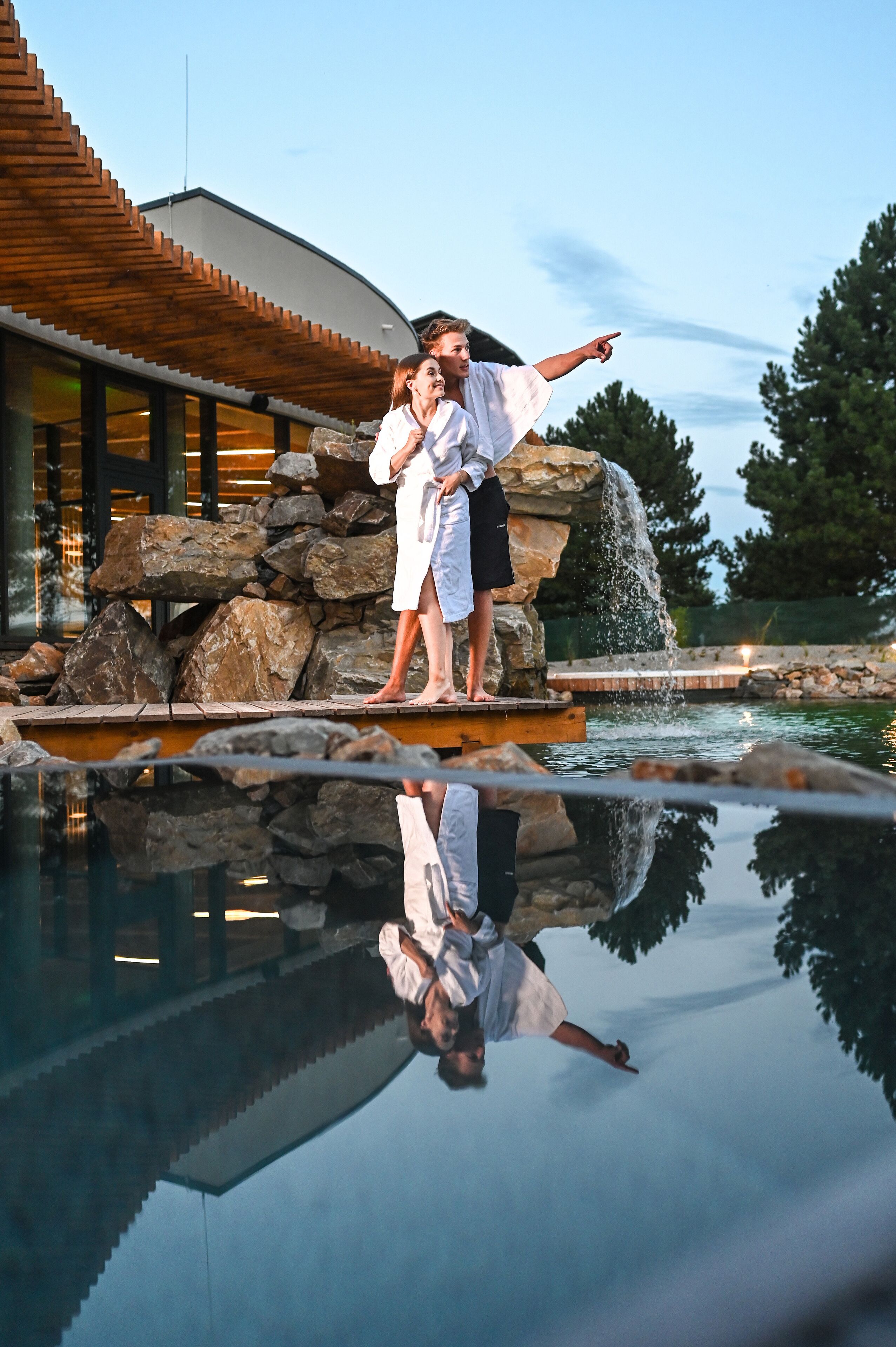 A couple standing on a wooden platform by a reflective pool, with a stone waterfall and a modern building in the background.