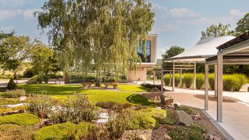Een modern gebouw met een glazen gevel, omgeven door een goed onderhouden tuin met diverse planten en een pergola.