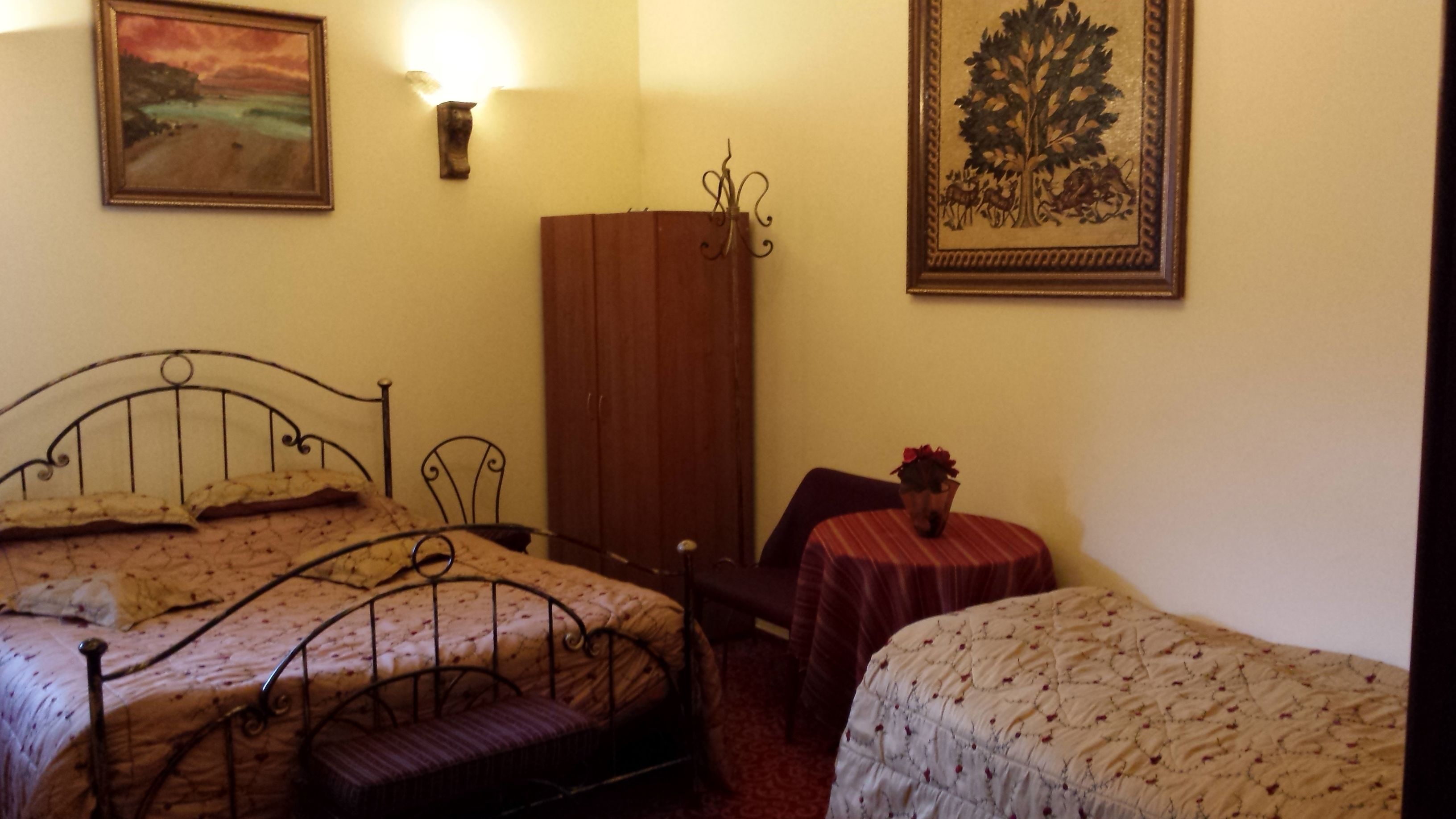 A room with two beds, a wooden wardrobe, a small table with a vase, and two wall-mounted lamps.