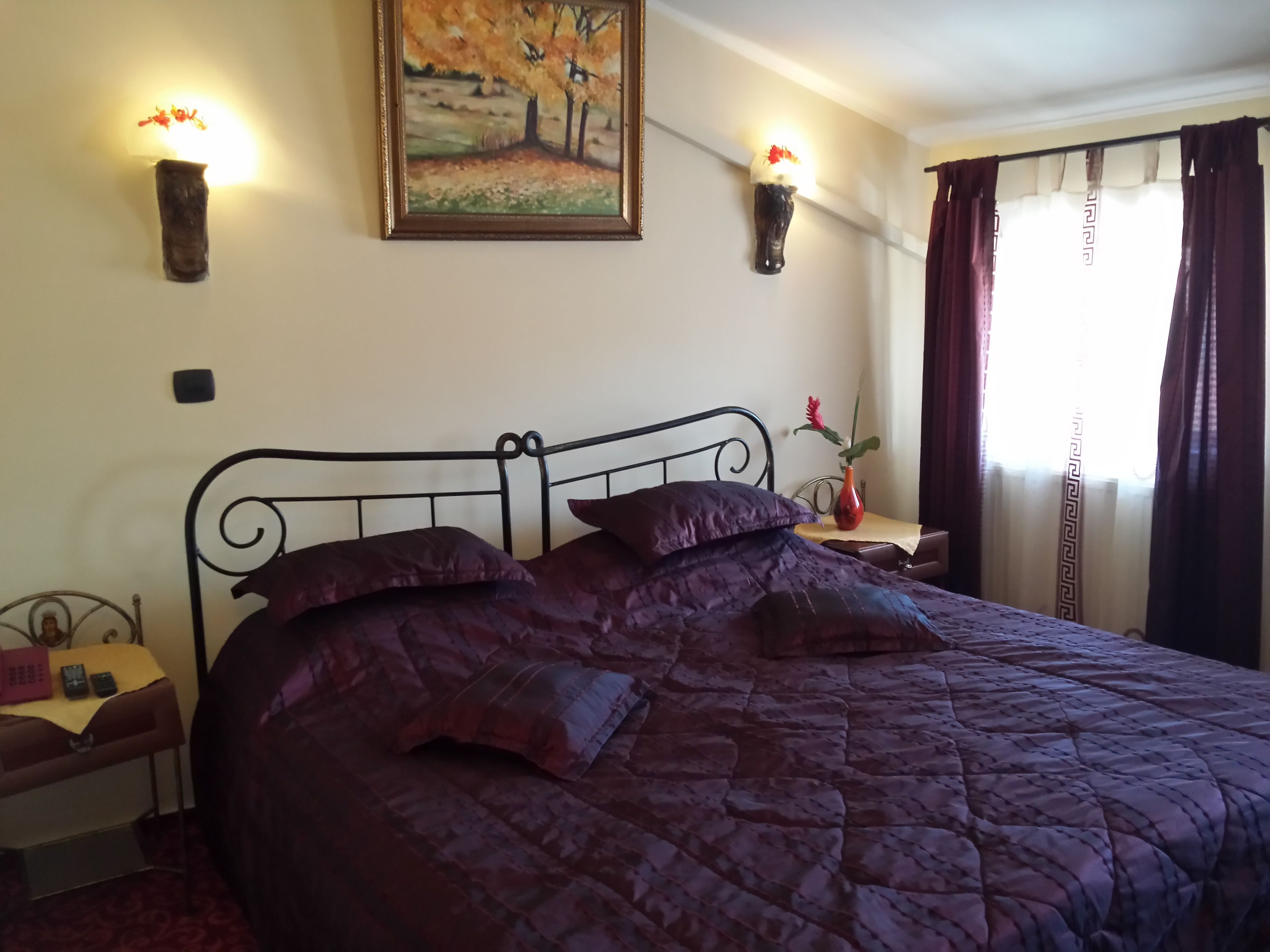 A bedroom with a bed, a nightstand, a wall-mounted lamp, a painting, and a window with curtains.
