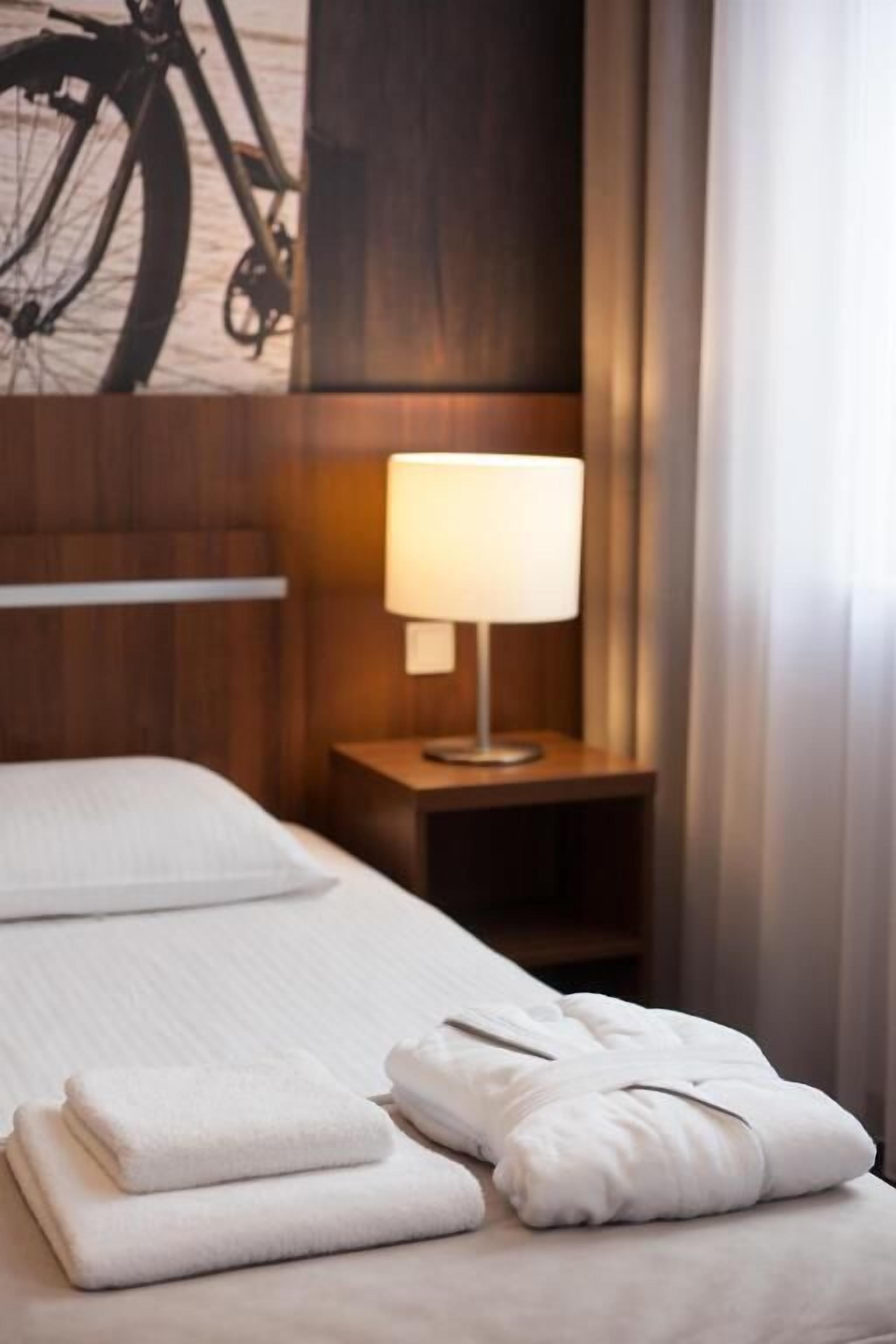 Siisti hotellihuone, jossa on huolitellusti pedattu sänky, yöpöydällä lamppu ja seinällä pyöräilyaiheinen taideteos.