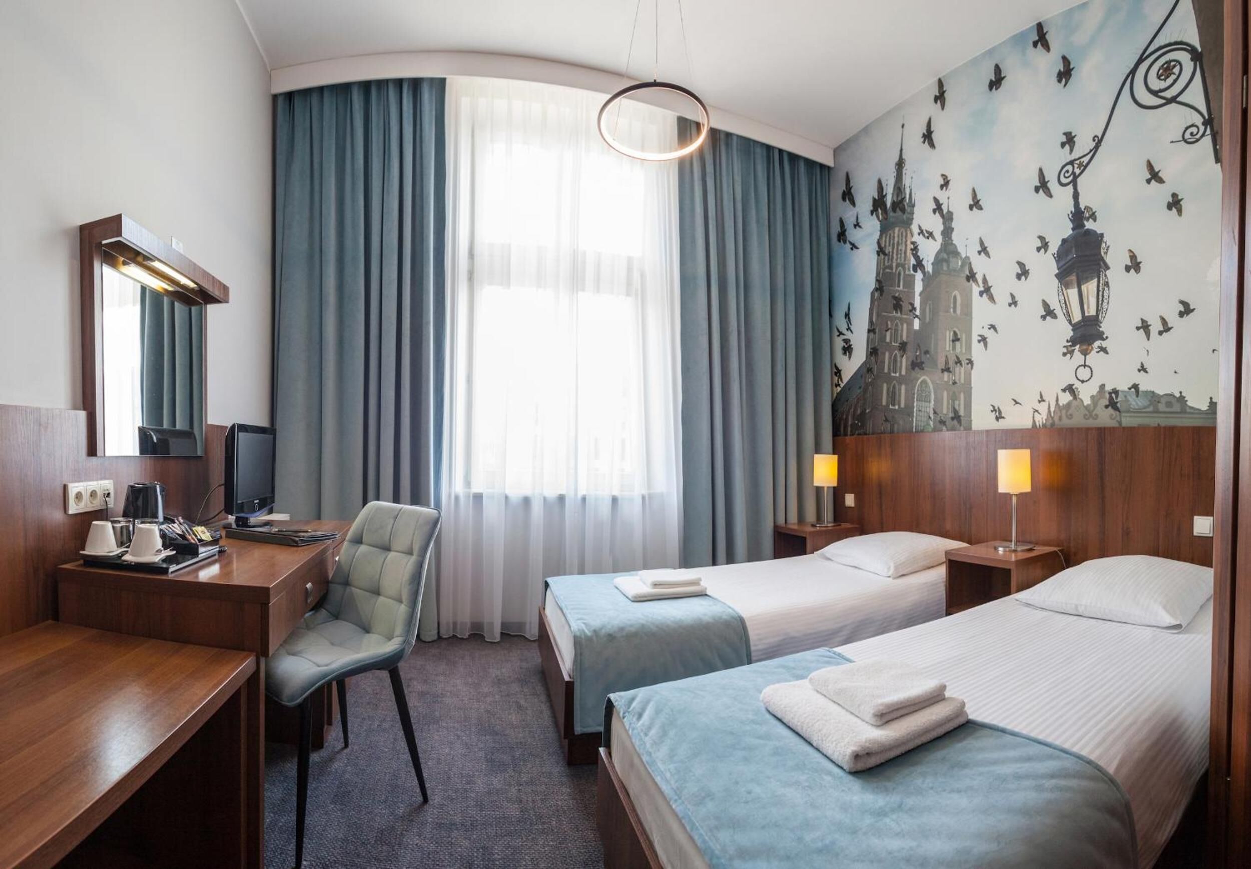 A hotel room with a desk, two beds, a chair, a TV, a lamp, and a large window with curtains.