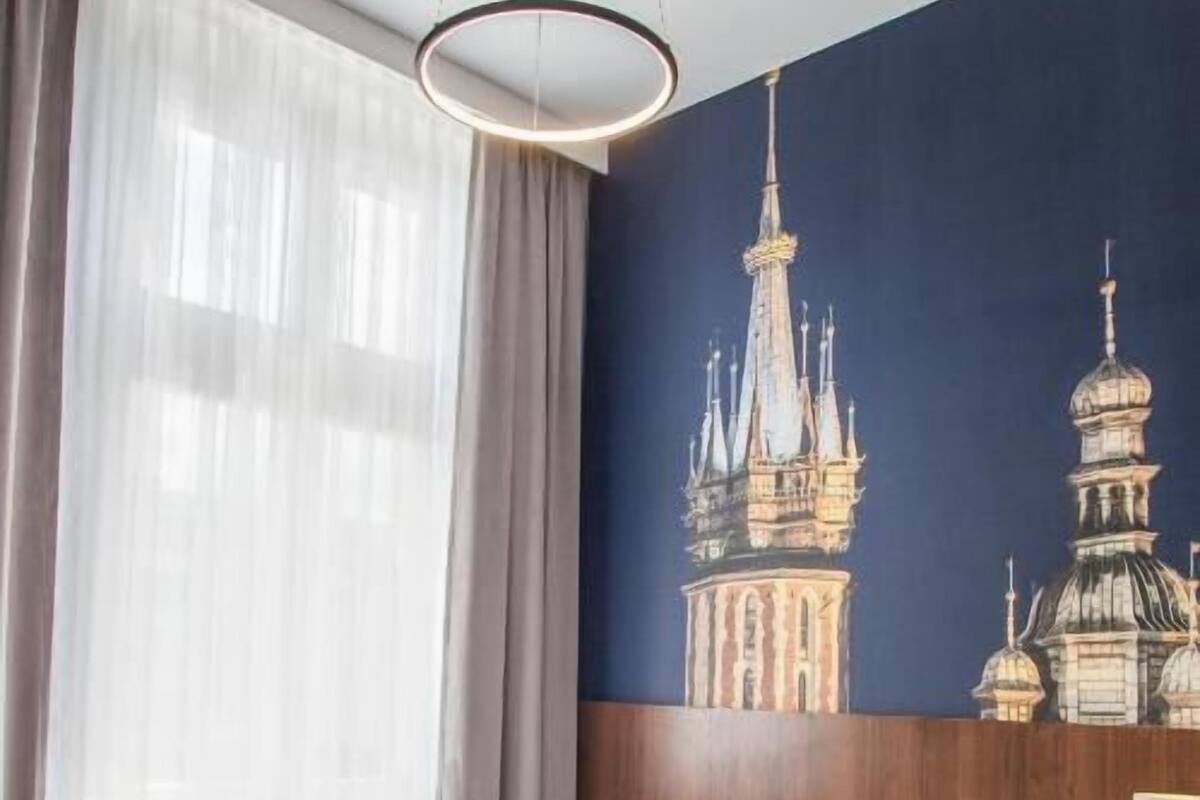 Quarto de hotel com cama, mesa de cabeceira, abajur e uma janela grande com cortinas leves.