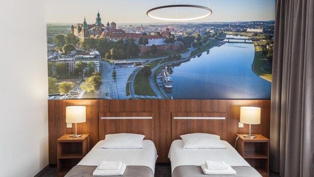 Une chambre d’hôtel avec deux lits, une grande fenêtre donnant sur un paysage urbain et une rivière, et un luminaire circulaire moderne au-dessus des lits.