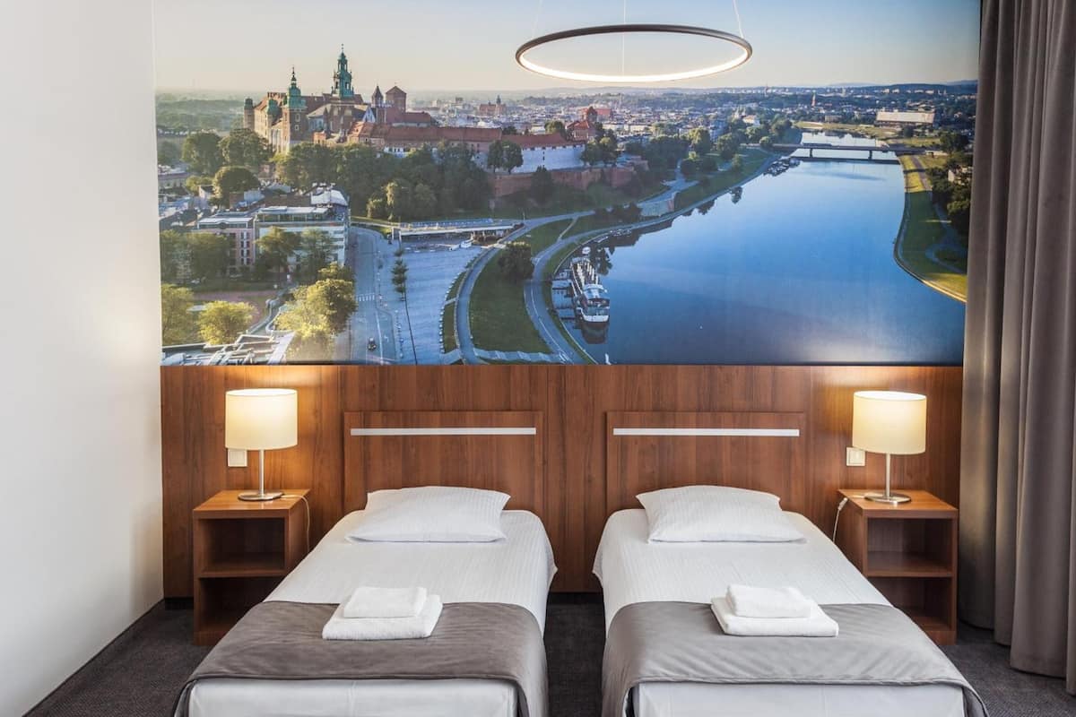 Quarto de hotel com duas camas, uma janela grande com vista para a paisagem urbana e o rio, e uma luminária circular moderna sobre as camas.