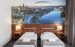Habitación de hotel con dos camas, un ventanal con vista a un paisaje urbano y un río, y una lámpara circular moderna sobre las camas.