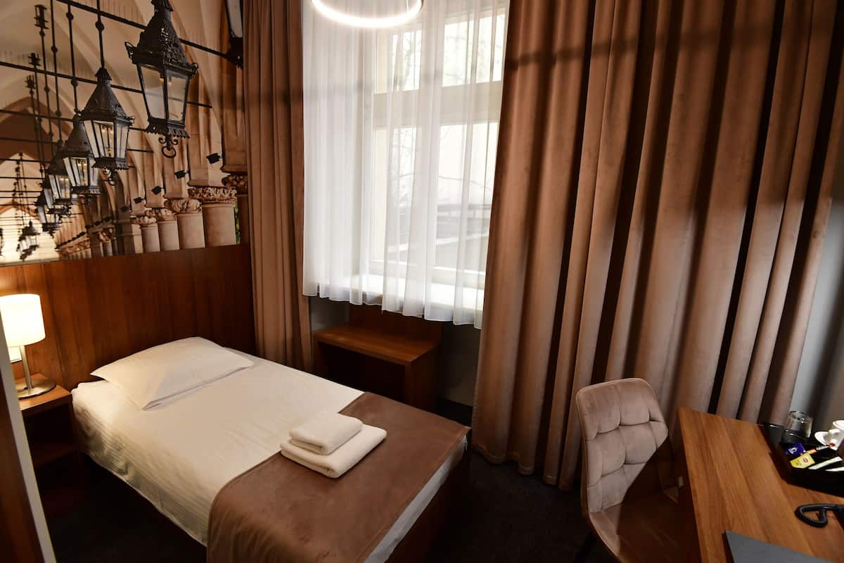 Quarto de hotel com cama bem arrumada, escrivaninha de madeira, uma cadeira e janela com cortinas.