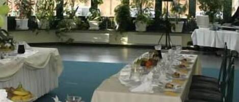 A restaurant with a long table set for a meal, surrounded by potted plants and a view of buildings.