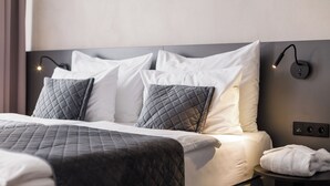 Una cama bien hecha con cabecera acolchada, almohadas blancas y grises, y una mesita de noche con una toalla enrollada.
