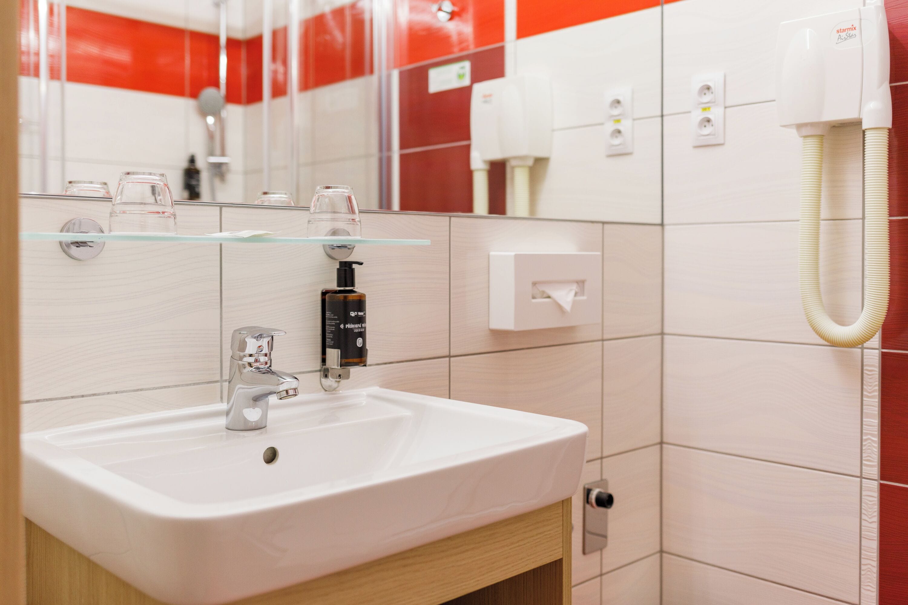Ein Badezimmer mit einem weißen Waschbecken, einer Glasablage, einem Spiegel und einer Wand mit roten und weißen Fliesen.