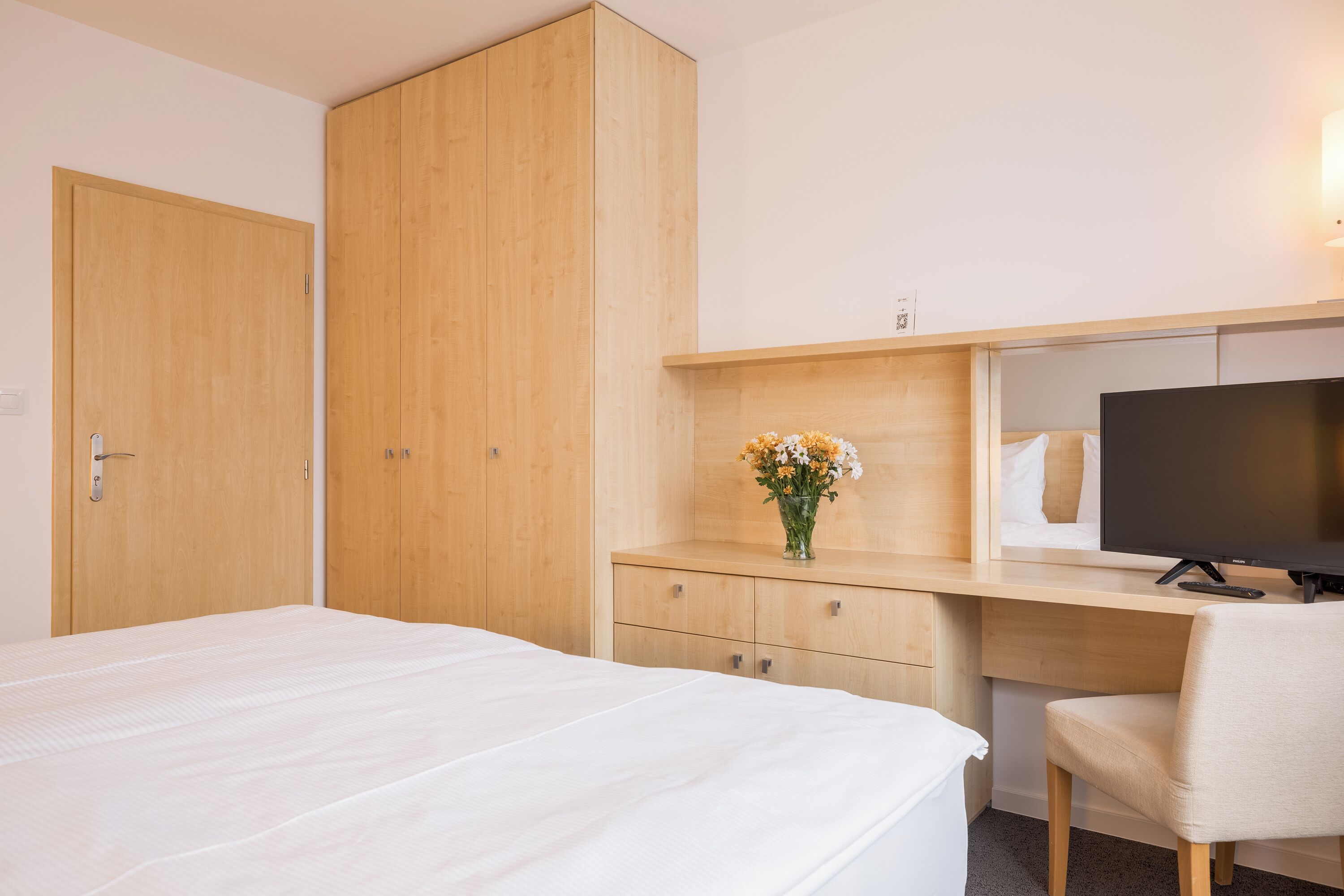 A hotel room with a bed, a desk with a TV, a chair, and wooden wardrobes.