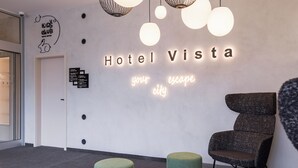 Un moderno vestíbulo de hotel con un letrero de neón que dice "Hotel Vista tu escape ciudad", un mostrador de recepción y una zona de estar con mesas y sillas redondas.