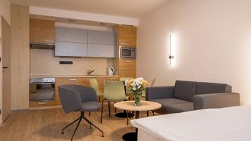 Una habitación de hotel moderna con kitchenette, un comedor con sillas, un sofá, una pequeña mesa redonda con un jarrón de flores y una cama.