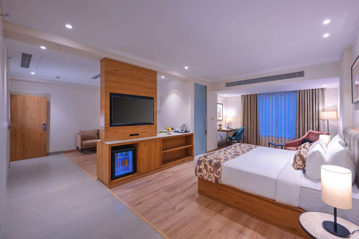 Une chambre d’hôtel moderne dotée d’un grand lit, d’une télévision à écran plat, d’un mini-réfrigérateur, d’un bureau avec une chaise et d’un coin salon comprenant une chaise et une table.
