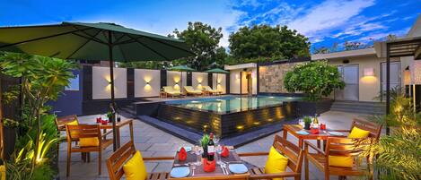 An outdoor dining area with tables and chairs, a pool, and surrounding greenery.