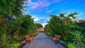 A pathway flanked by lush greenery and wooden benches, leading to a building entrance. - Royale Sarovar Portico Agra (Agra)