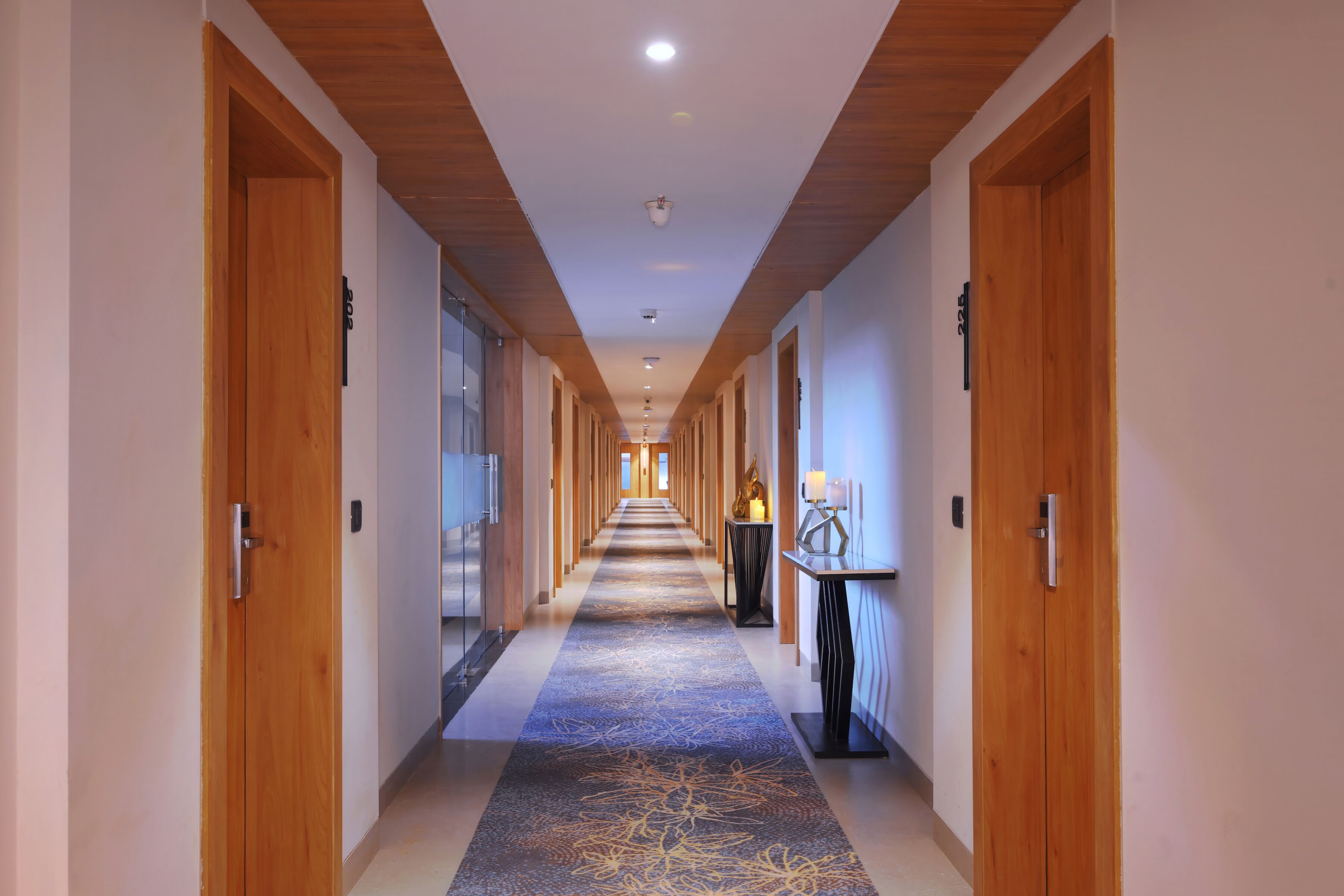 Un couloir d’hôtel avec des portes en bois, une moquette bleue à motifs et un long couloir menant à une chambre équipée d’une lampe et d’une petite table.
