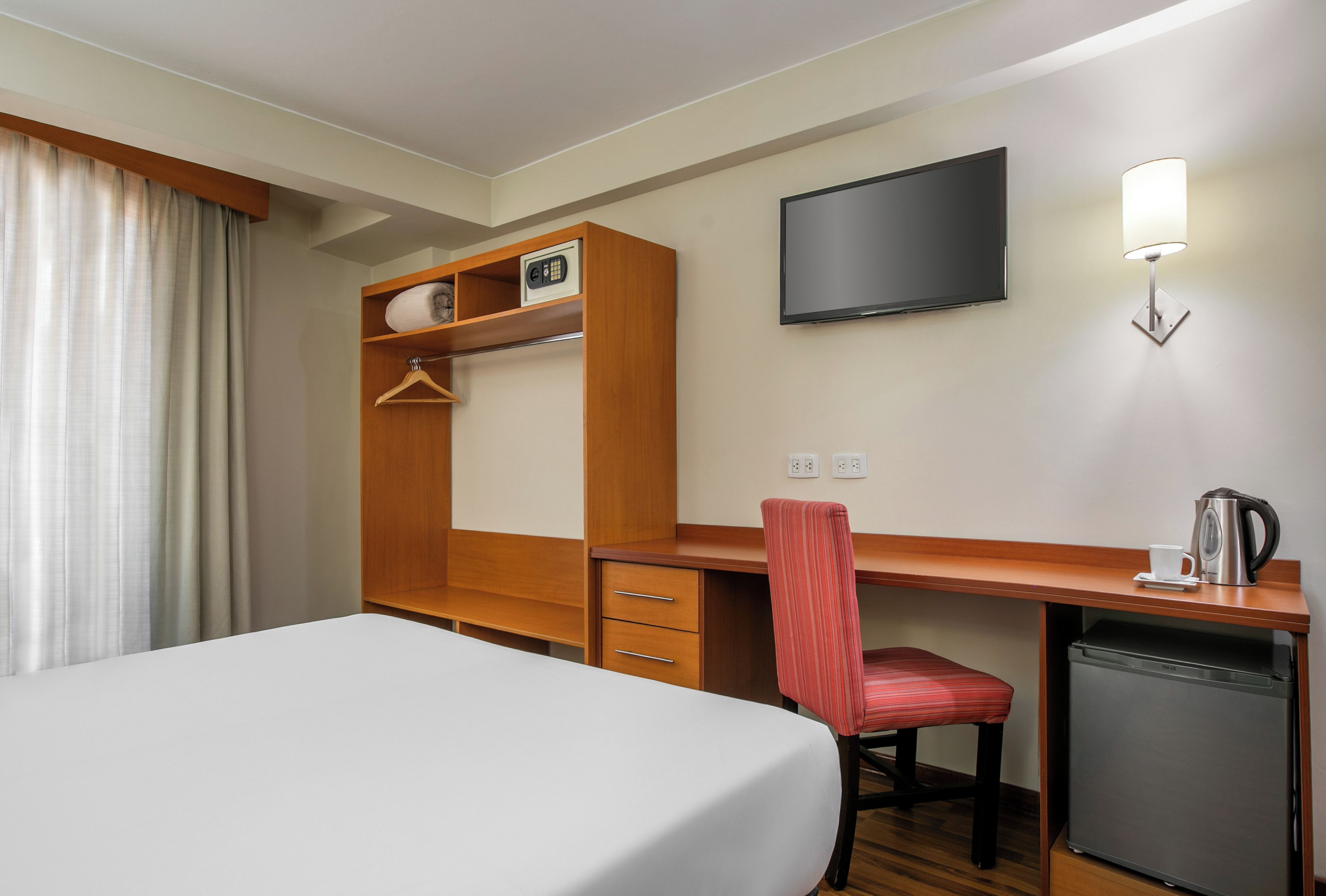 A hotel room with a bed, a desk with a chair, a television, a kettle, and a small refrigerator.