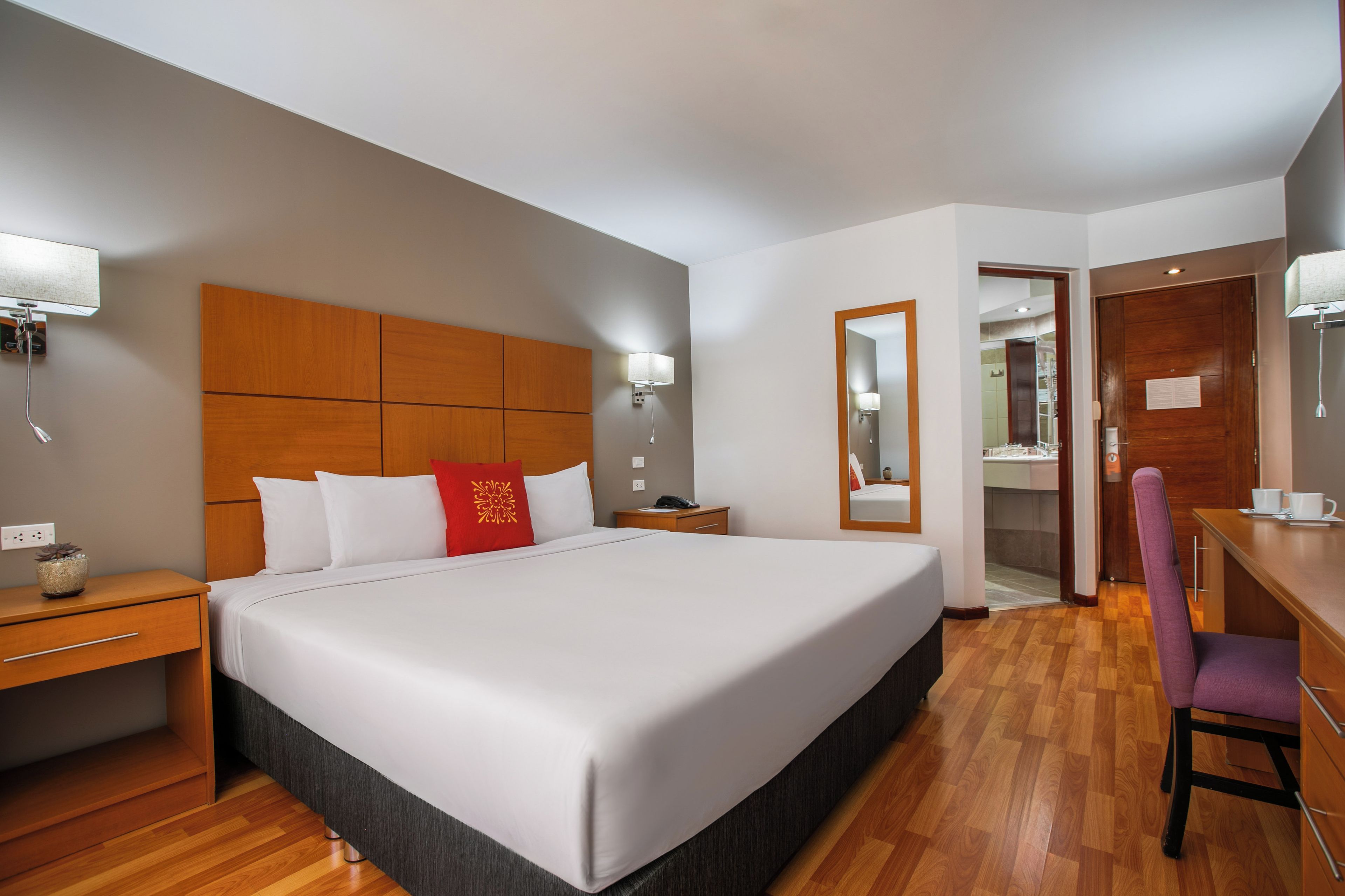 A hotel room with a large bed, bedside tables, a desk, a chair, a mirror, and a wardrobe.