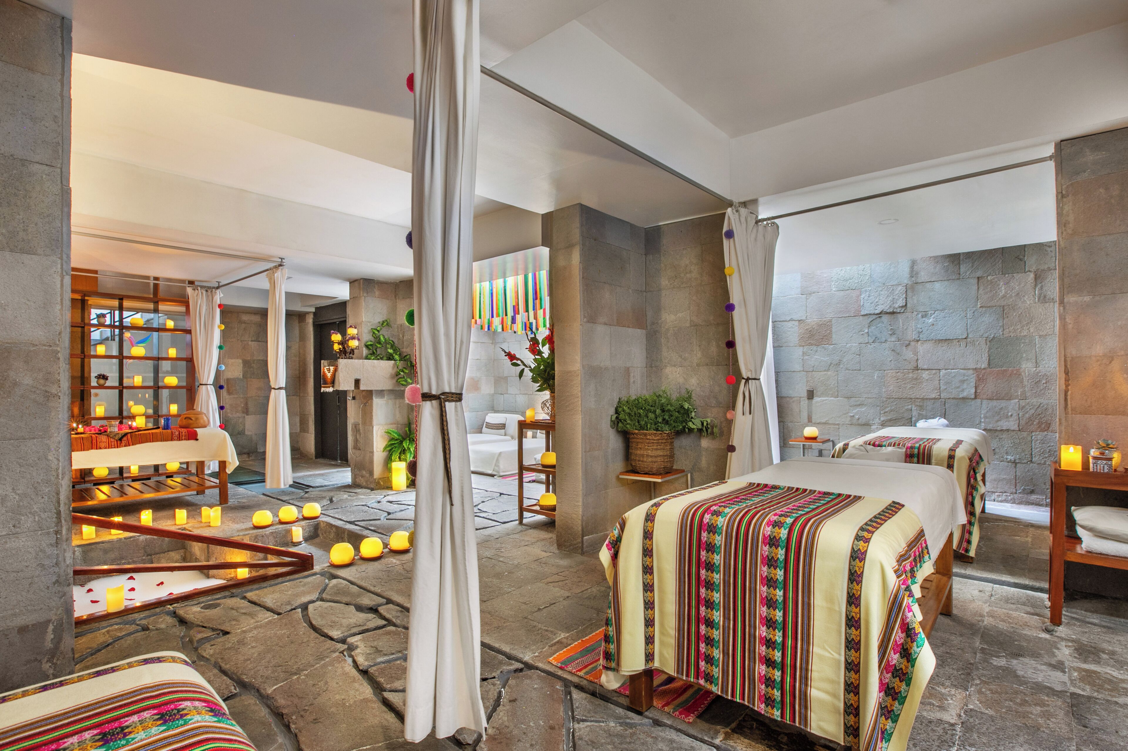 Habitación de hotel con pared de piedra, cama con manta colorida, zona de estar con mesa y sillas, y un pequeño balcón con plantas.
