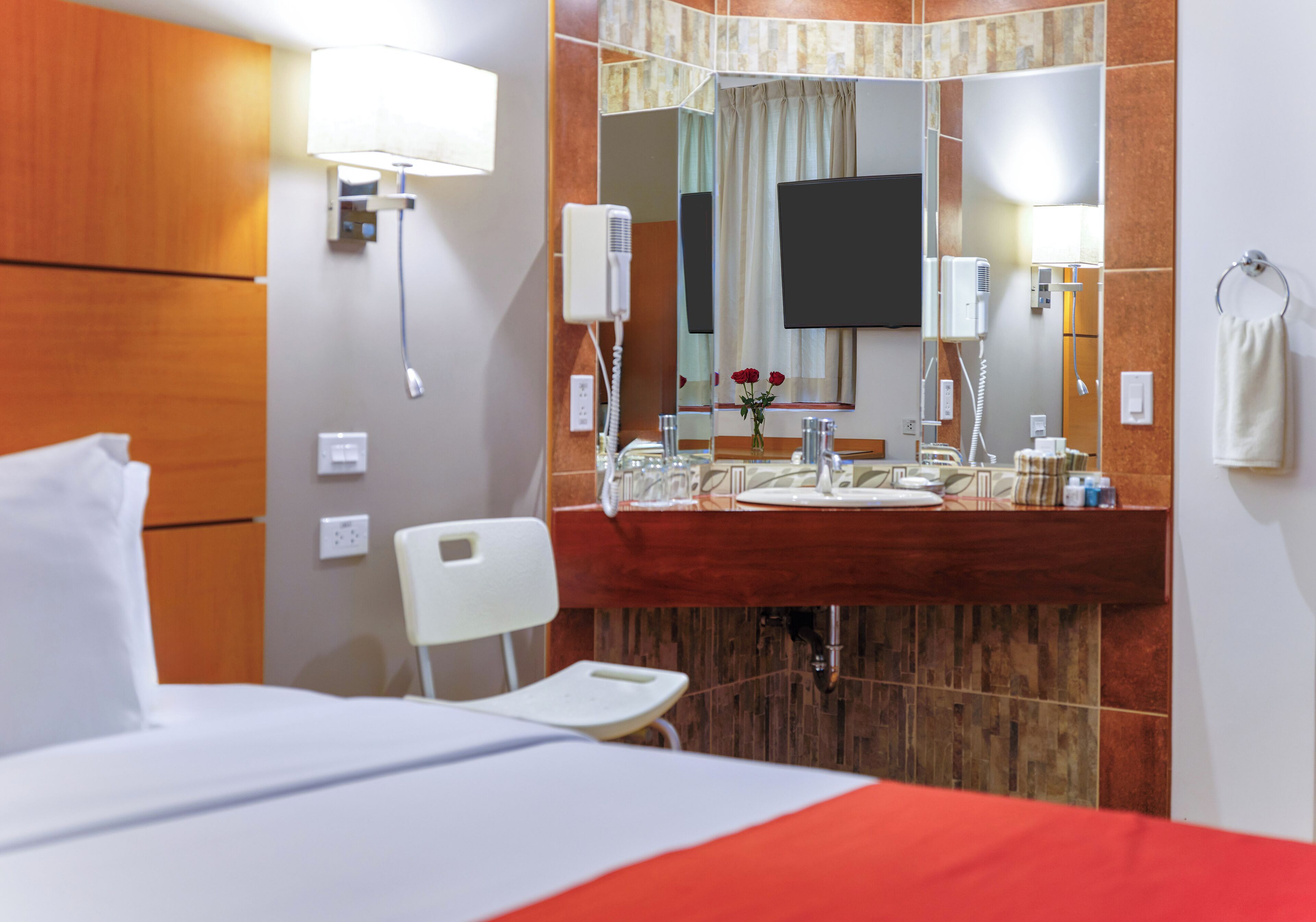 A hotel room with a bed, a wooden headboard, a large mirror, a vanity with a sink, and a television mounted on the wall.