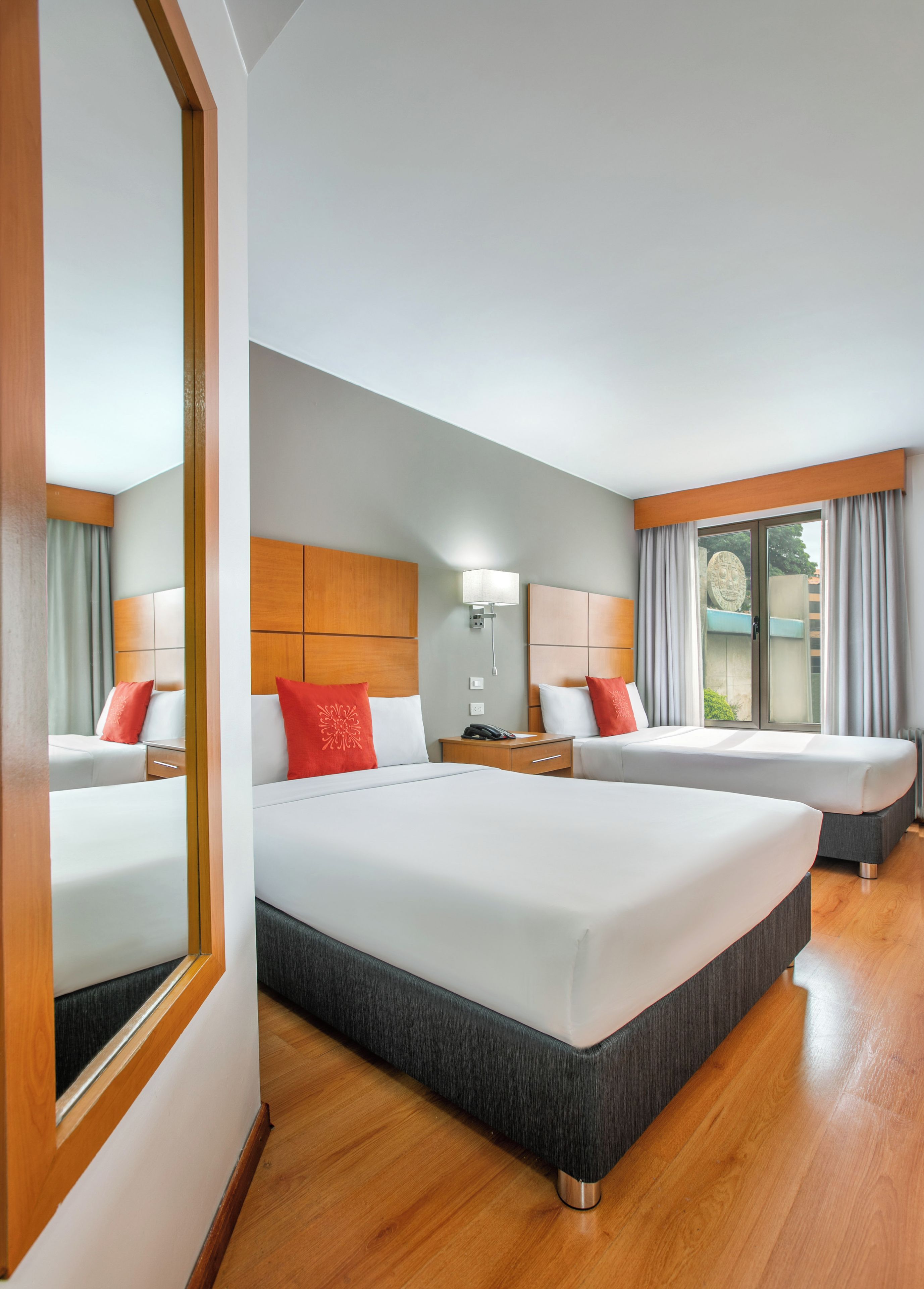 Habitación de hotel con dos camas, suelo de madera, mesita de noche con lámpara y vistas por la ventana.