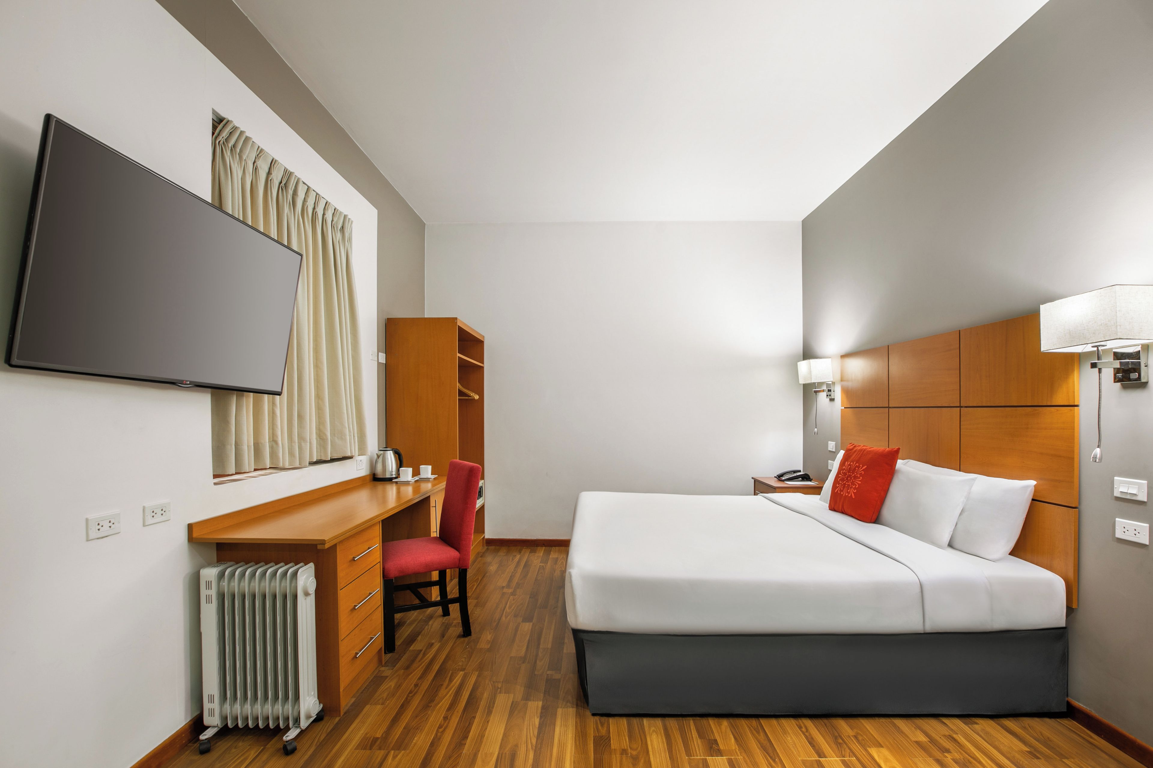 A hotel room with a bed, a desk, a chair, a radiator, and a television.