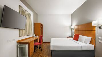 A hotel room with a bed, a desk, a chair, a radiator, and a television.