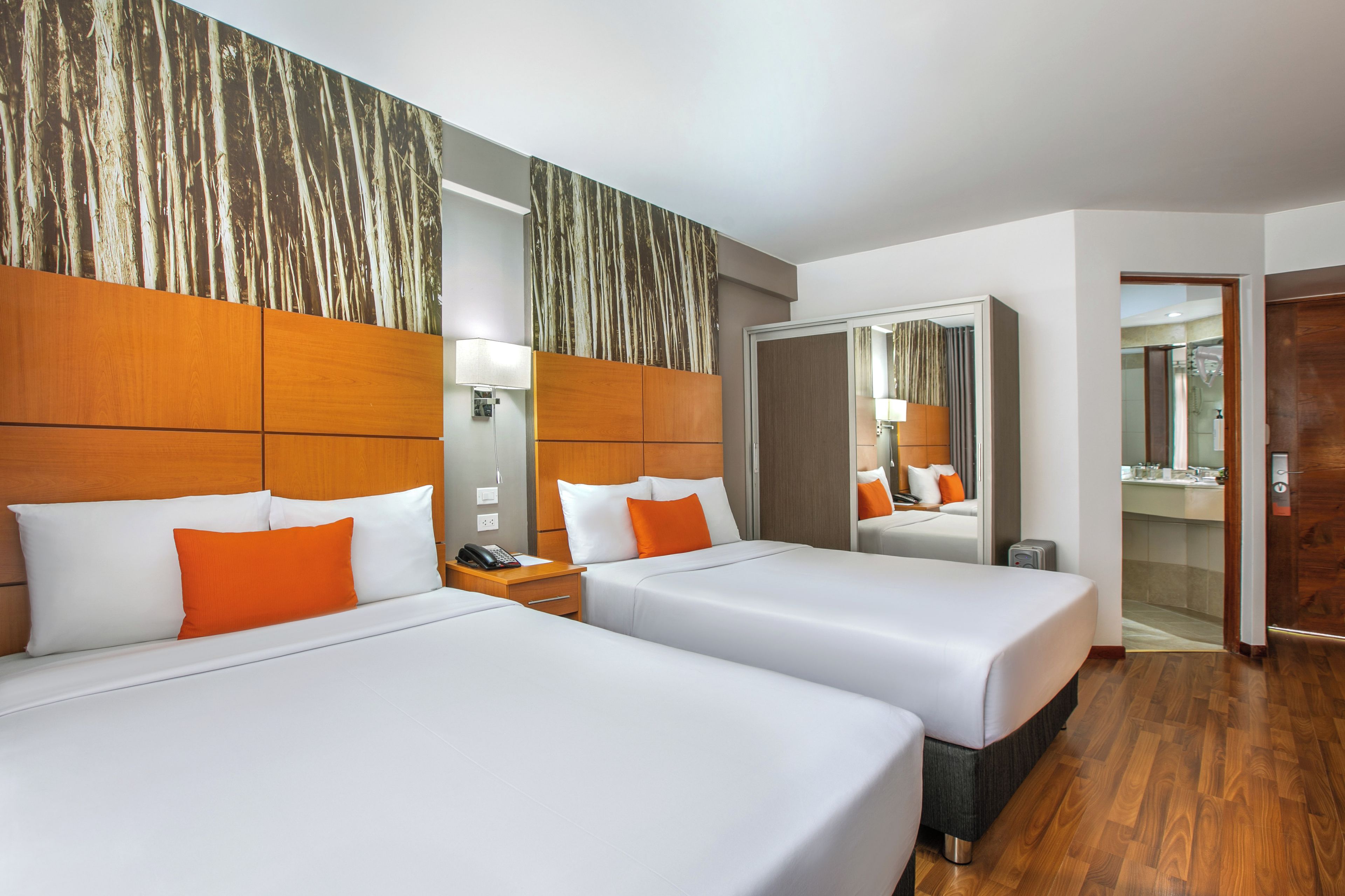 A hotel room with two beds, a wooden headboard, a mirror, a nightstand, and a bathroom visible through an open door.