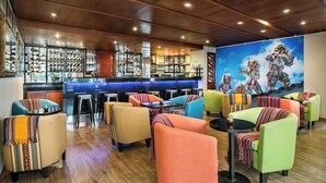 Zona de bar con diversas opciones de asientos, incluyendo taburetes y sillas acolchadas, una barra con botellas y vasos, y un mural que representa figuras tradicionales.