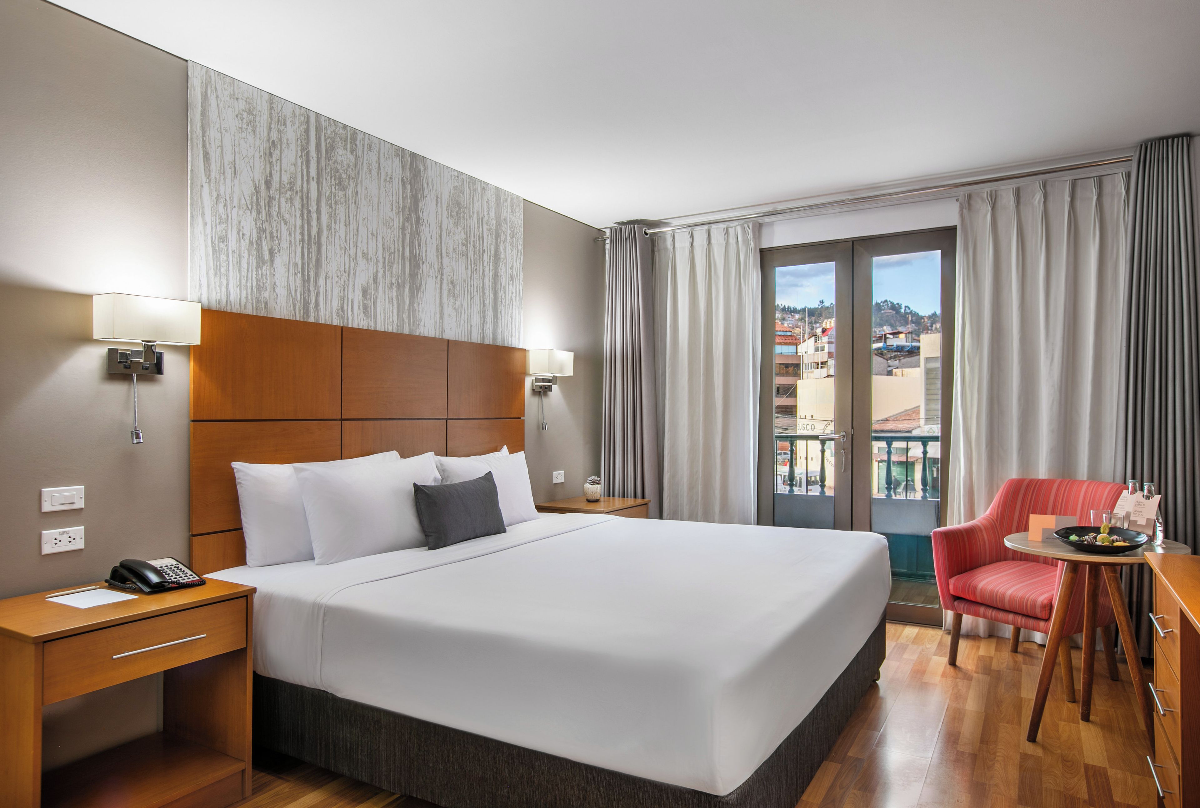 A hotel room with a large bed, a desk with a telephone, a chair, and a view of a cityscape through the window.