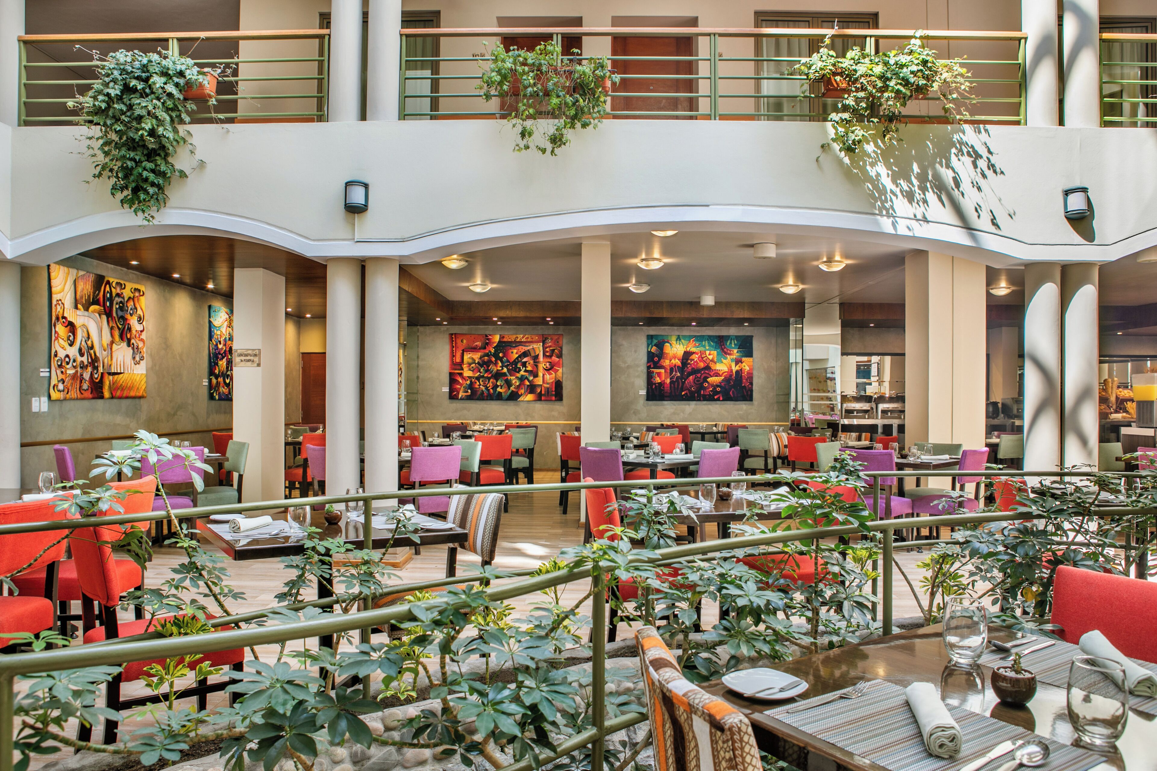 A spacious indoor dining area with multiple tables and chairs, surrounded by potted plants and artwork on the walls.