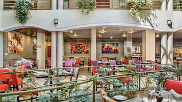 A spacious indoor dining area with multiple tables and chairs, surrounded by potted plants and artwork on the walls.