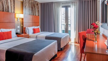 Quarto de hotel com duas camas, uma escrivaninha, uma cadeira, um abajur e uma janela com cortinas.