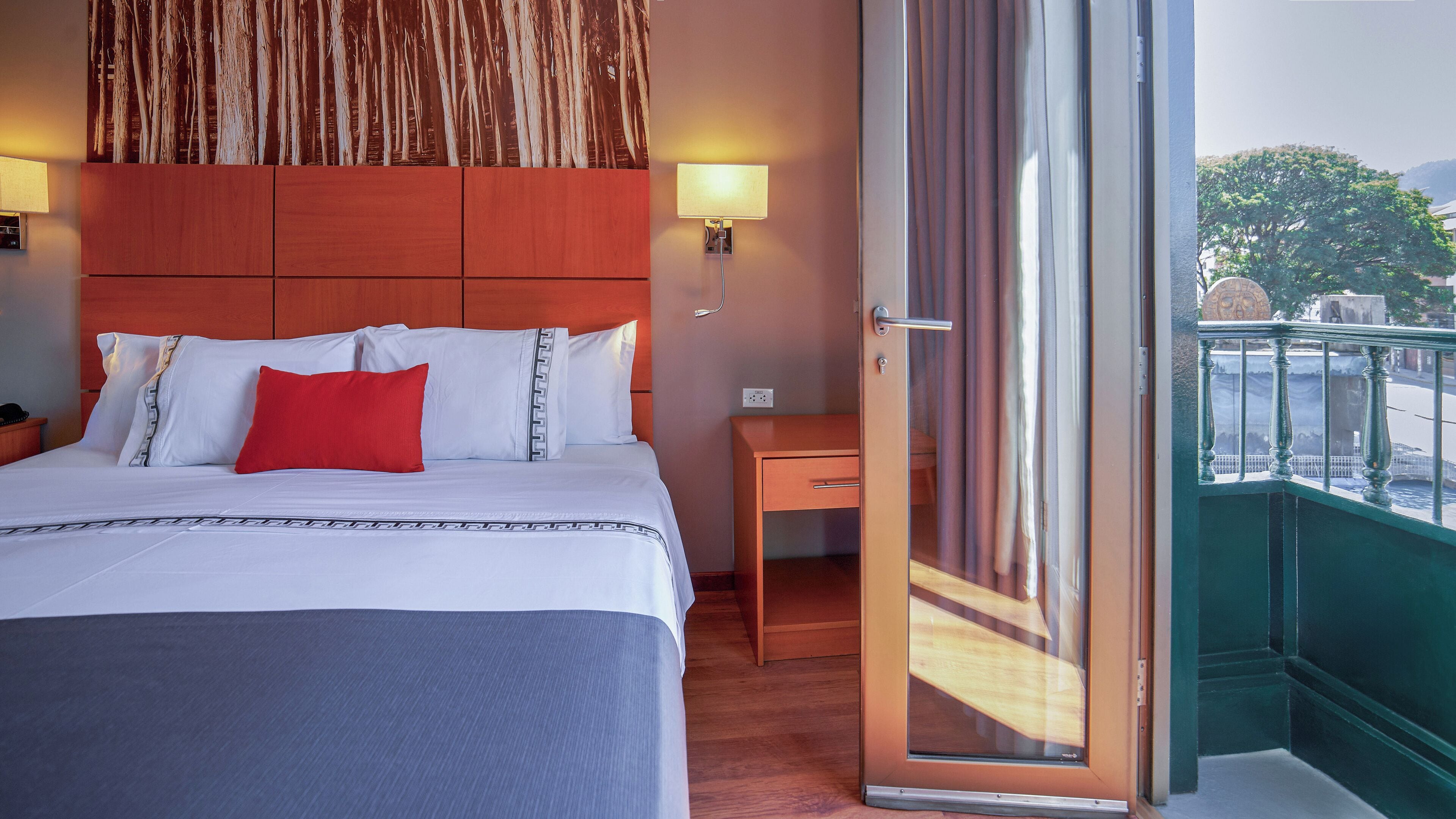 A hotel room with a bed, bedside table, and a view of the outside balcony.