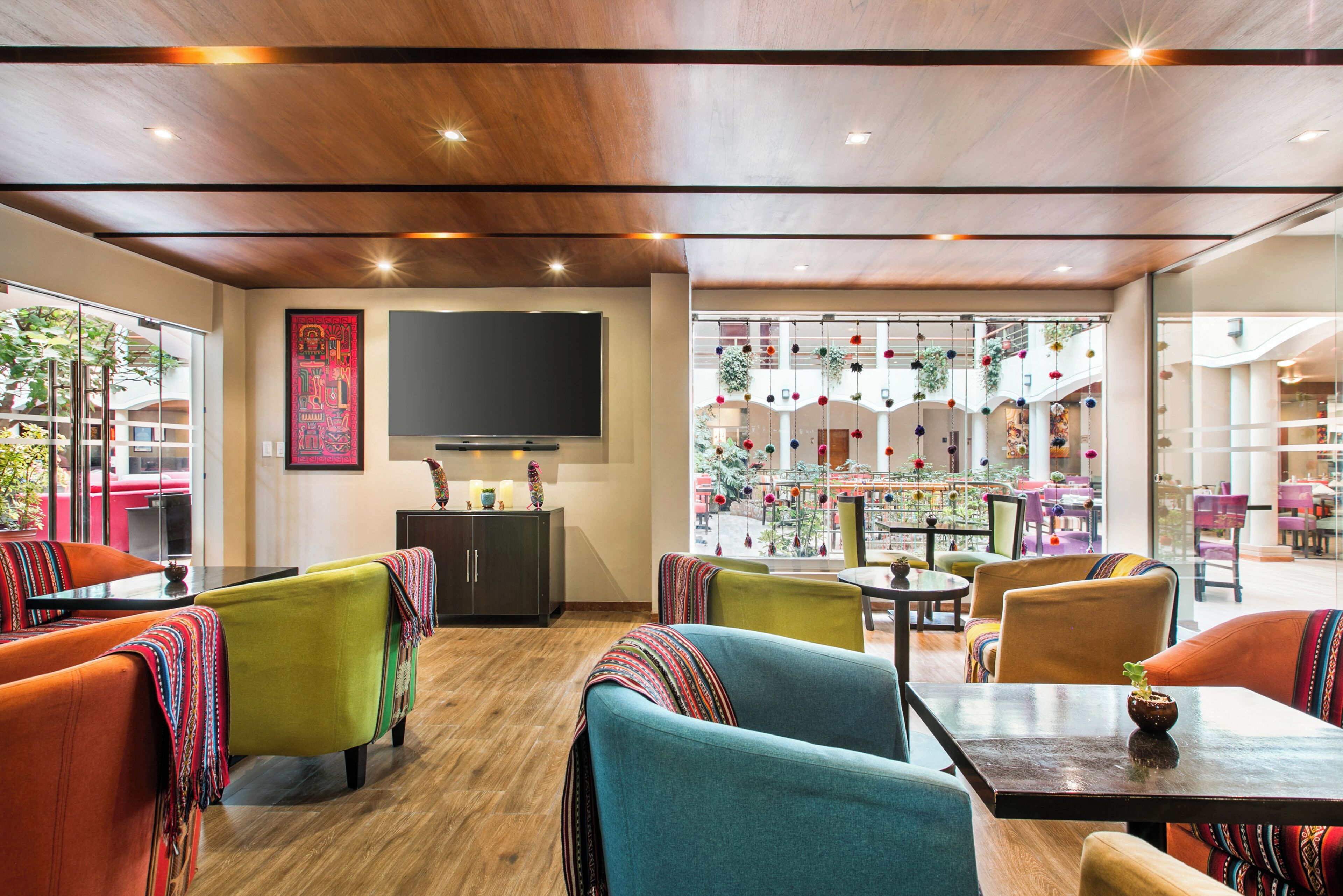 A modern interior with wooden ceiling, a flat-screen TV, colorful seating, and a view of an outdoor dining area.