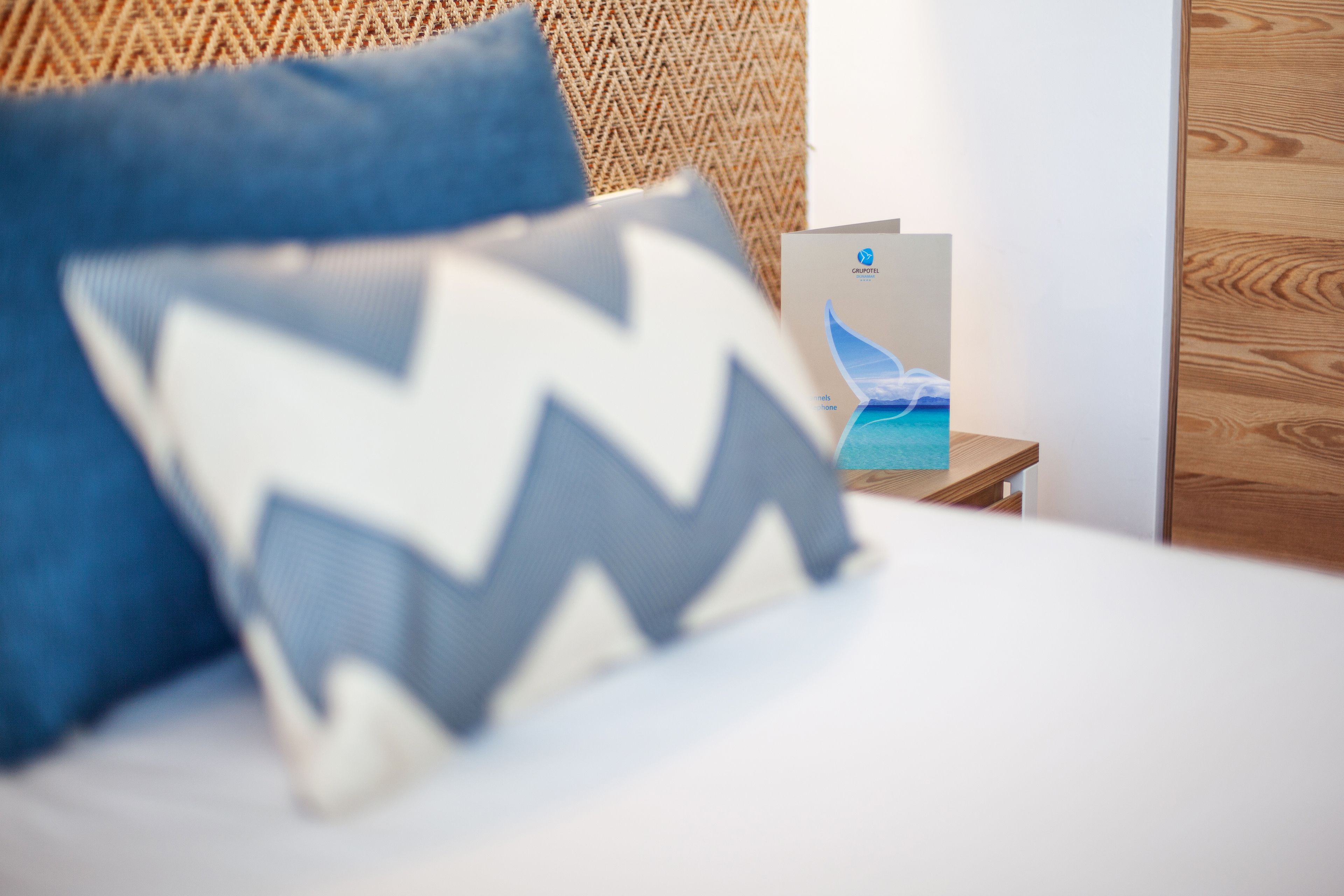 Una cama con un cojín azul y blanco, una pared de madera con textura y una mesita con un folleto.