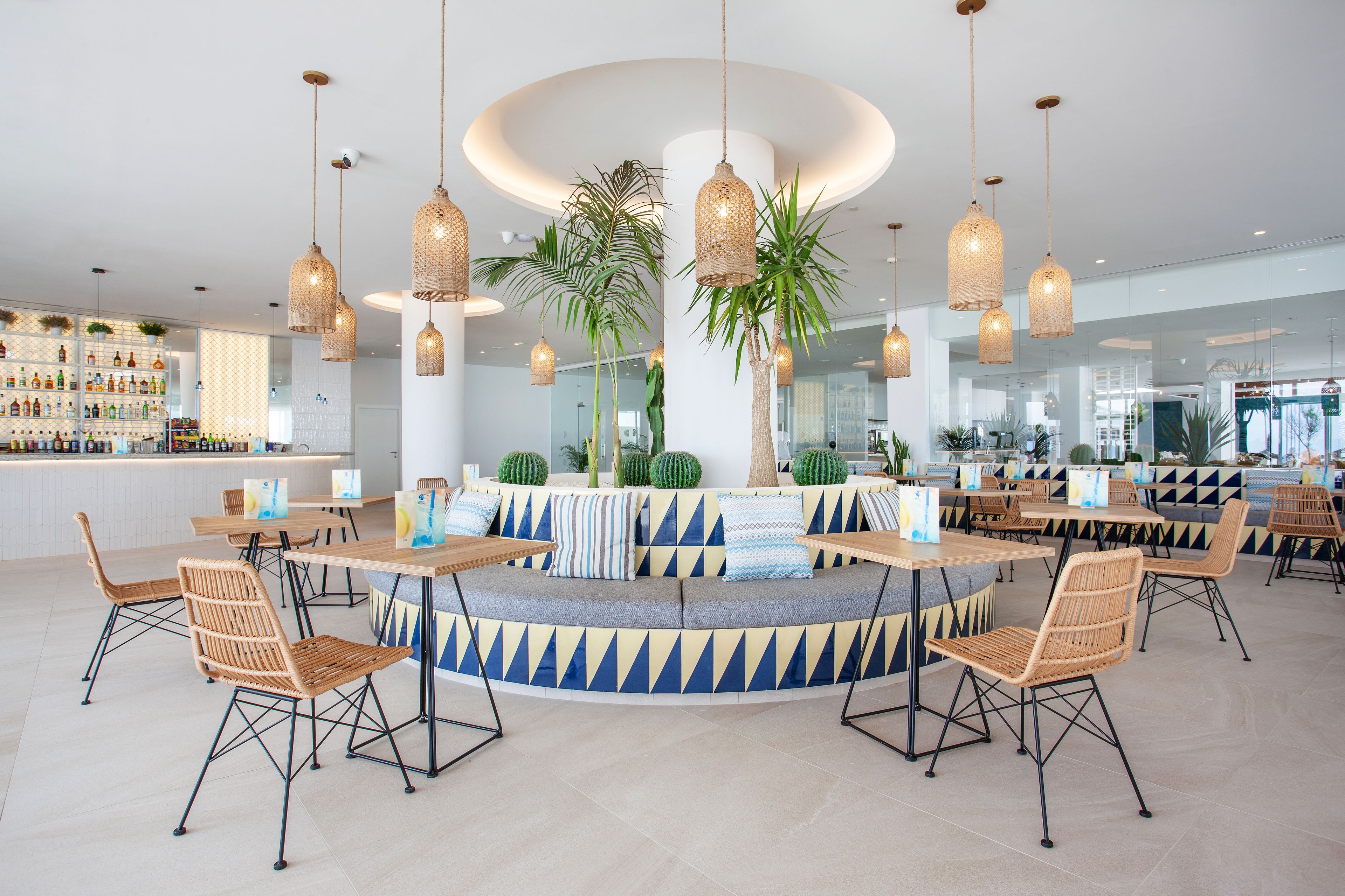 A modern lounge area with a central seating arrangement, hanging wicker lamps, and a bar with a variety of bottles.