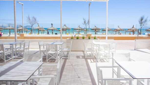 Un área de estar al aire libre con mesas y sillas blancas, con vista a una playa con sombrillas y el mar.
