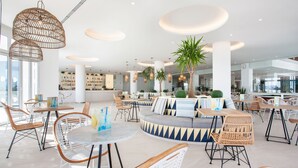Un espacio interior moderno con mesas redondas, sillas de mimbre y una zona de estar central con un sofá y cojines.