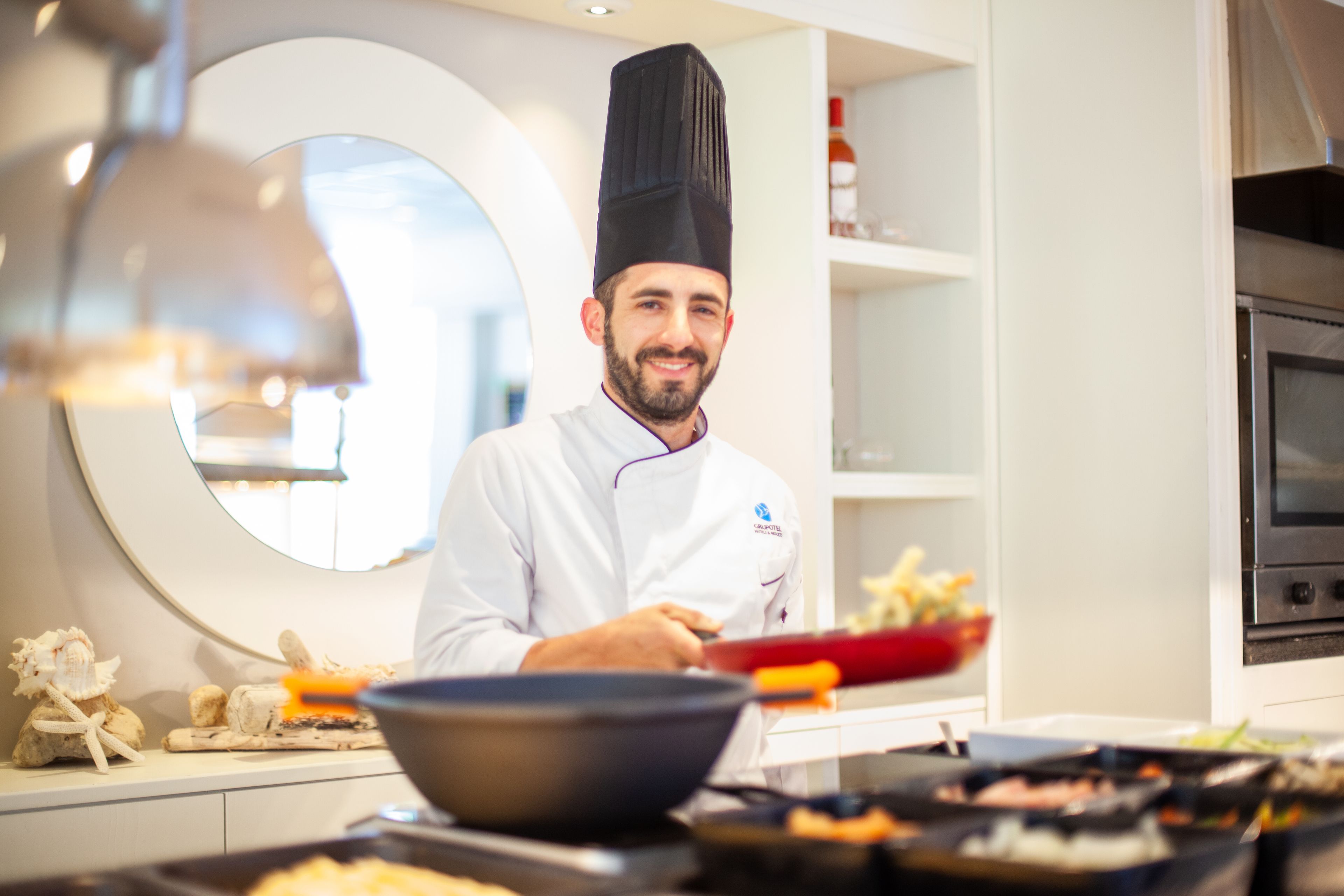 Un chef vêtu d’un uniforme blanc et coiffé d’un haut-de-forme noir se tient dans une cuisine moderne équipée d’un miroir rond, d’une cuisinière et de divers ustensiles de cuisine.