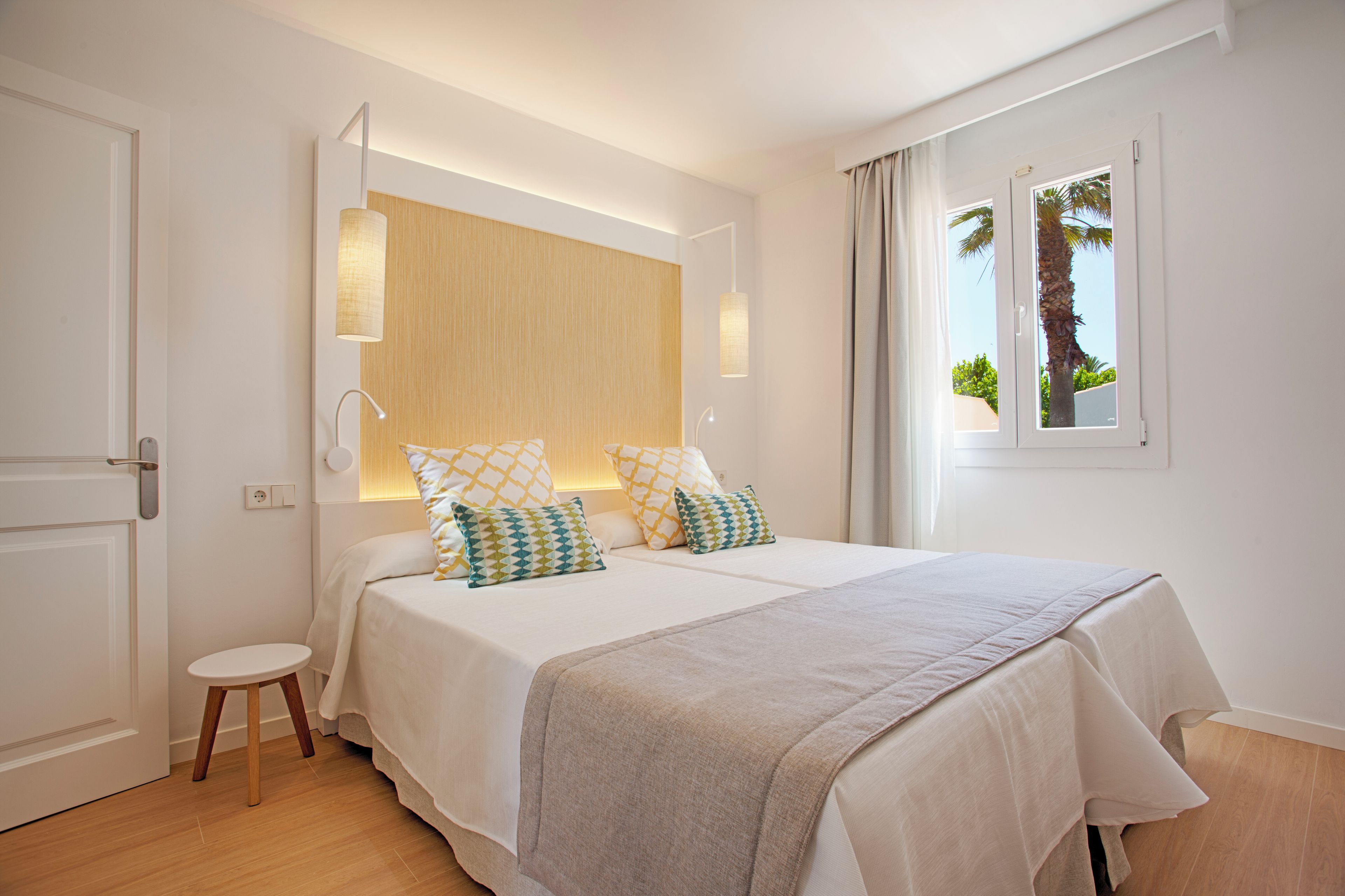 Habitación de hotel moderna con una cama grande, un taburete pequeño y una ventana que ofrece vistas a palmeras.