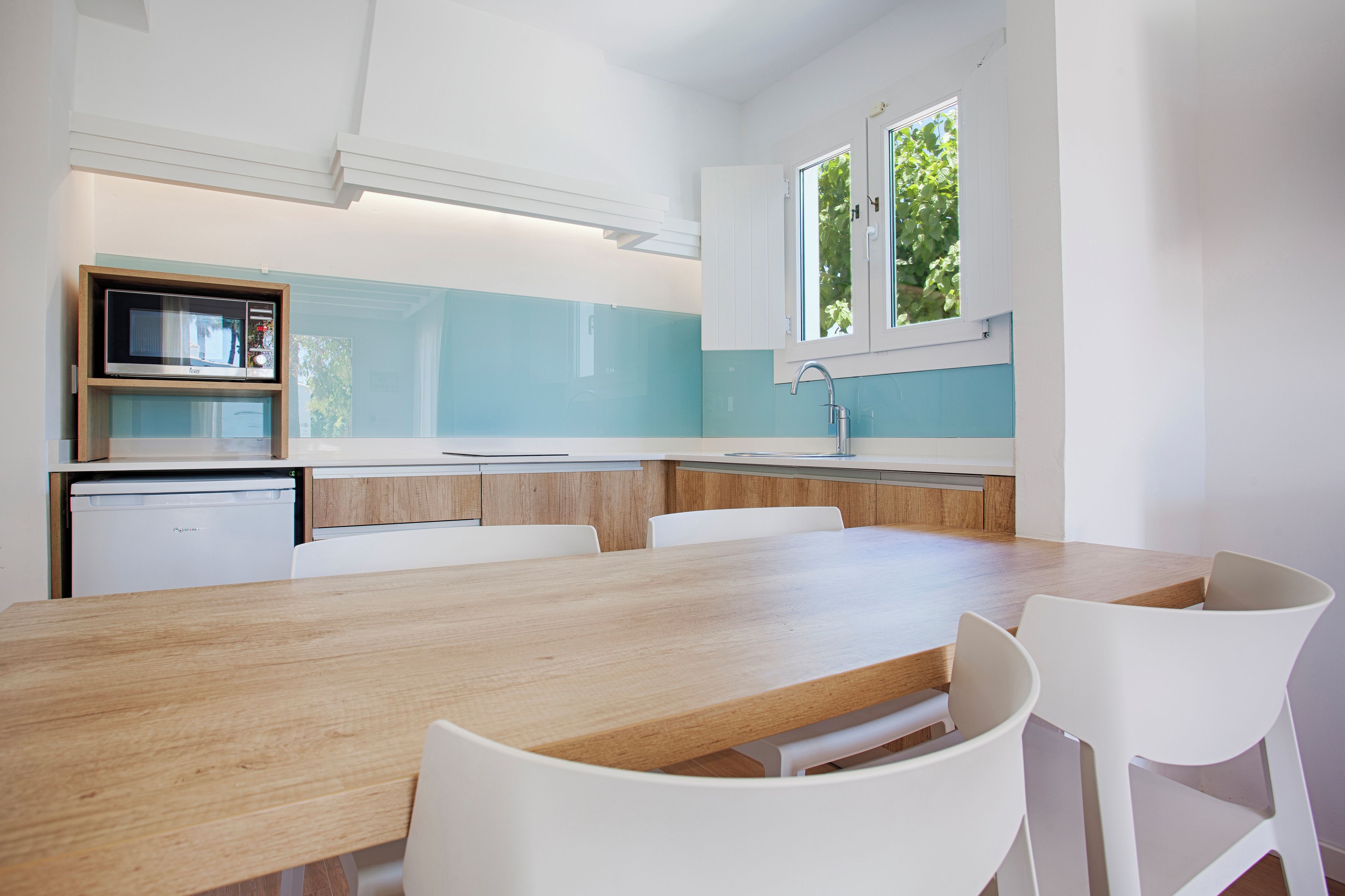 Une cuisine moderne avec une table en bois et des chaises blanches, un four encastré et une fenêtre donnant sur un paysage verdoyant.