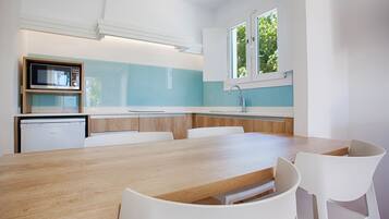 A modern kitchen with a wooden table and white chairs, a built-in oven, and a window with a view of greenery.