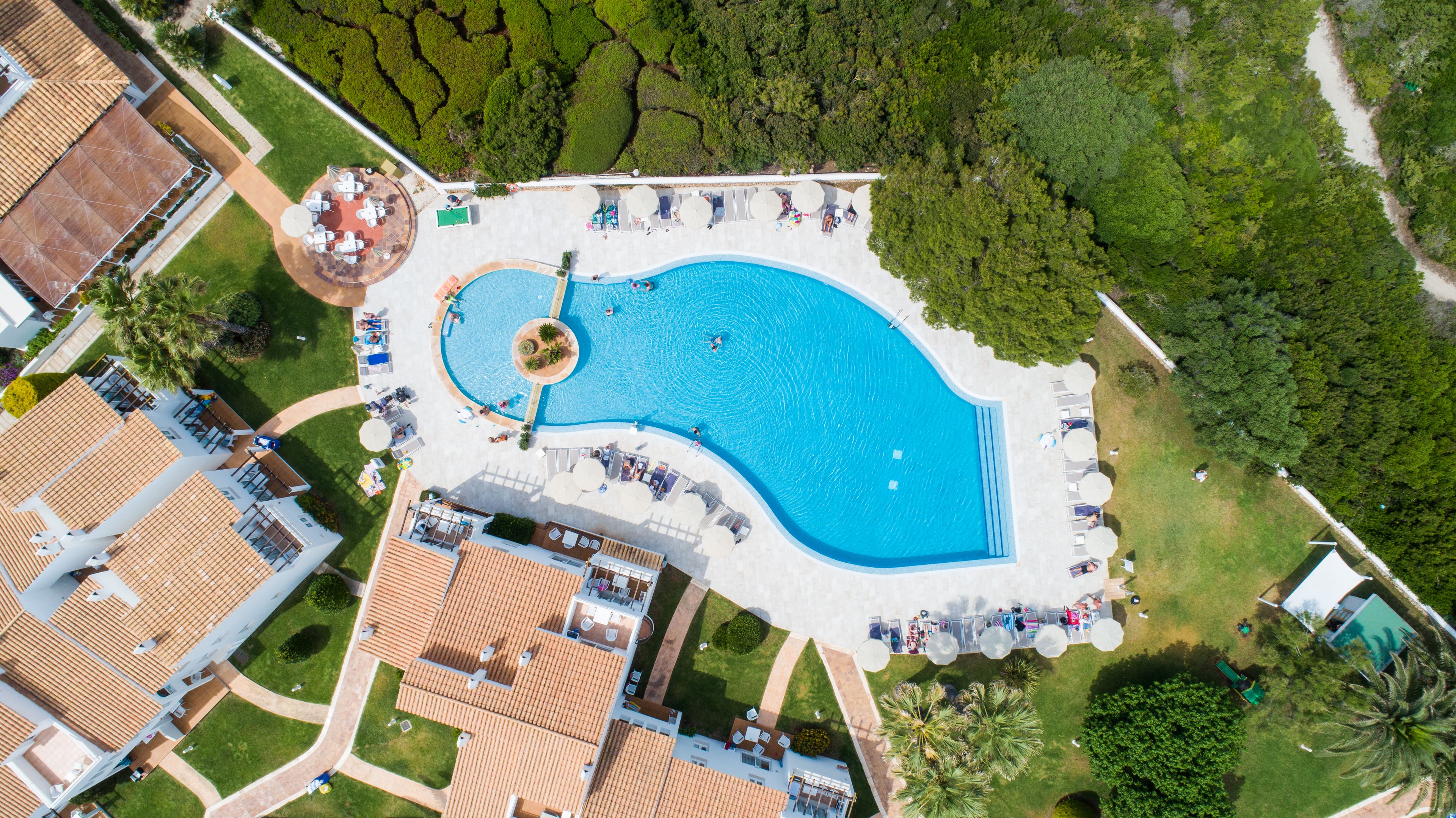 An aerial view of a resort with a large swimming pool, multiple lounge areas, and surrounding greenery.